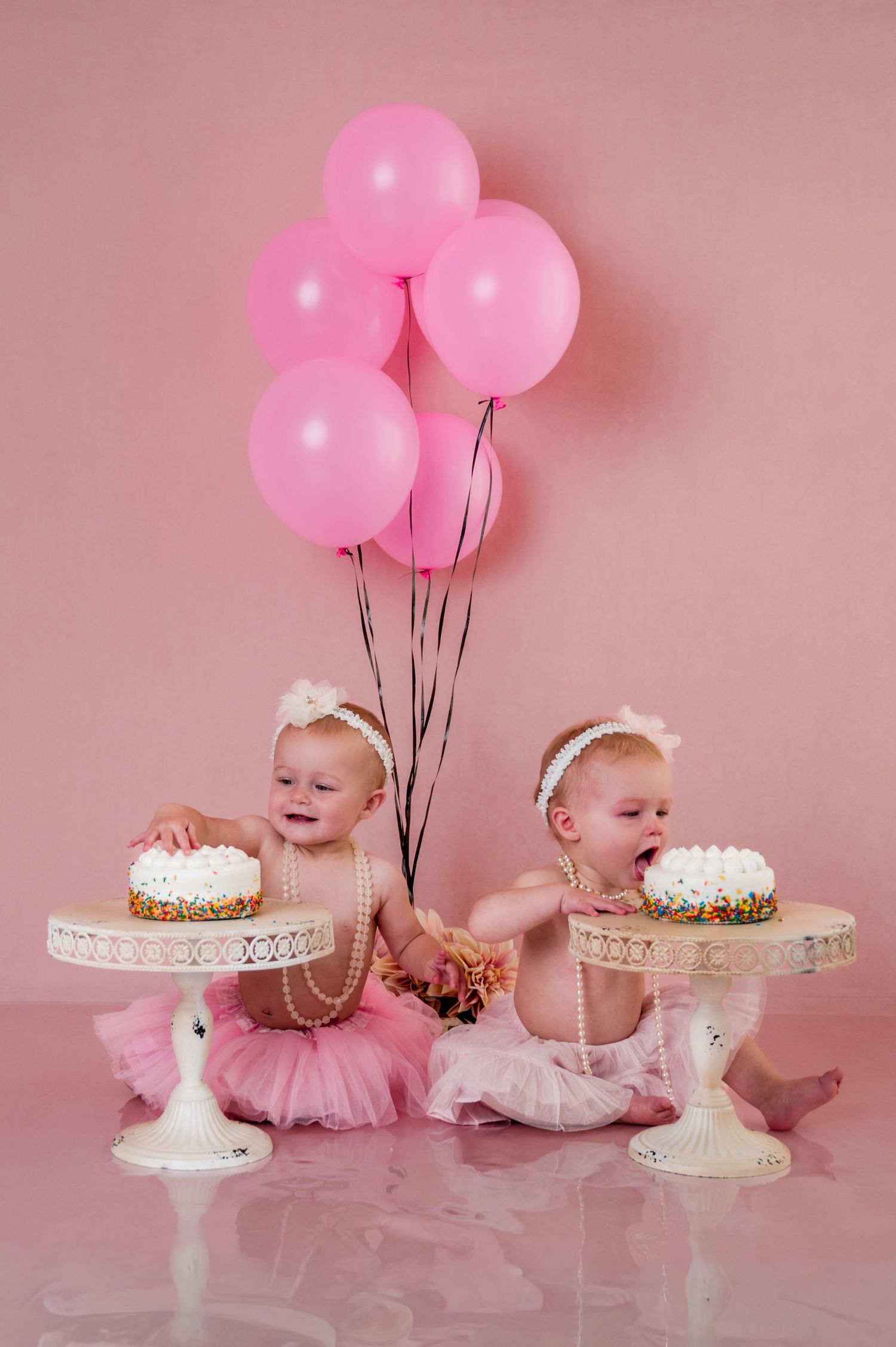 Two babies in pink and white tutus sit with birthday cakes and pink balloons against a rosy backdrop, Kewaskum, WI Studio Cake Smash