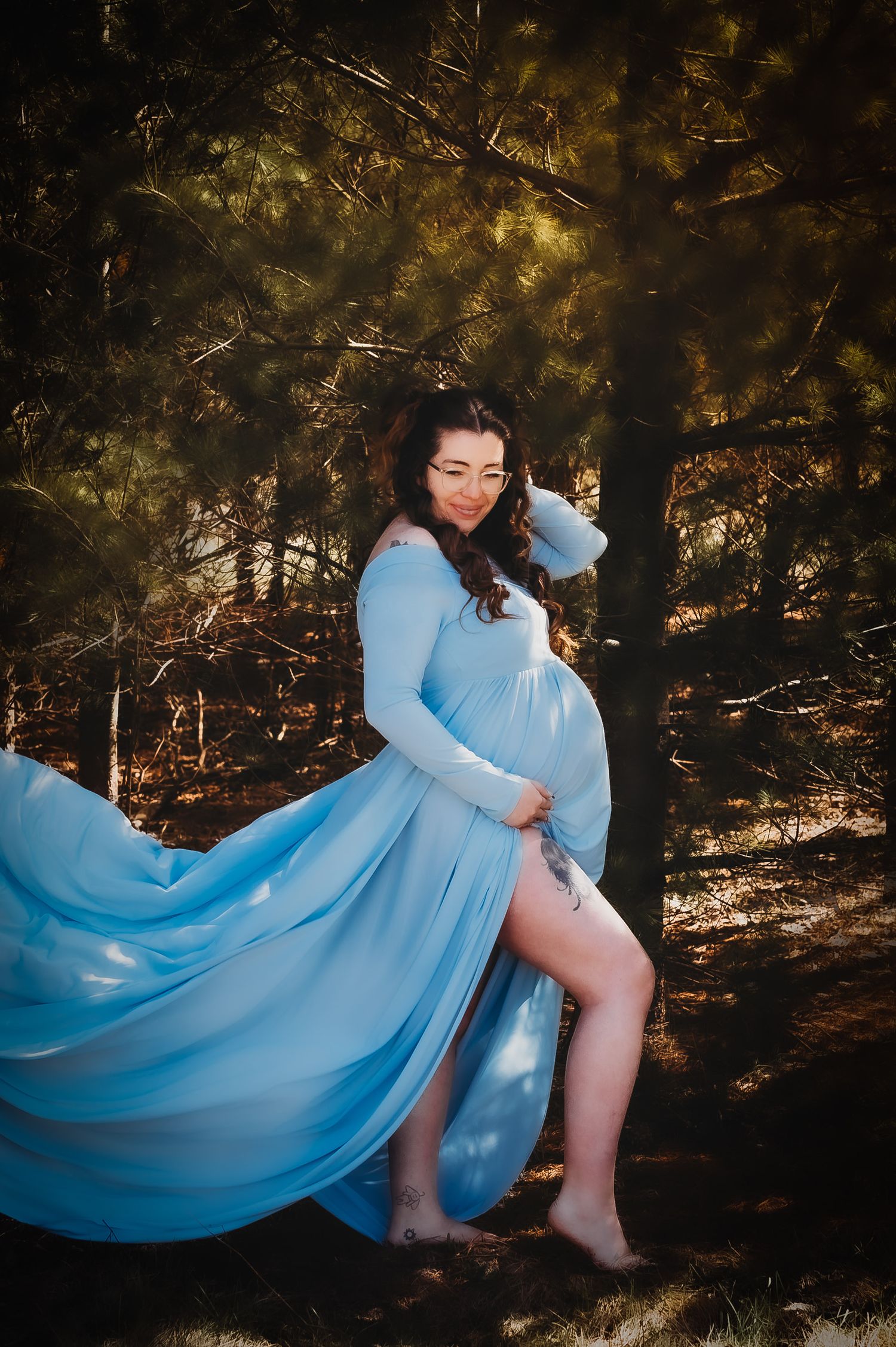 Ethereal maternity portrait in a blue gown with flowing fabric in a sunlit forest setting.