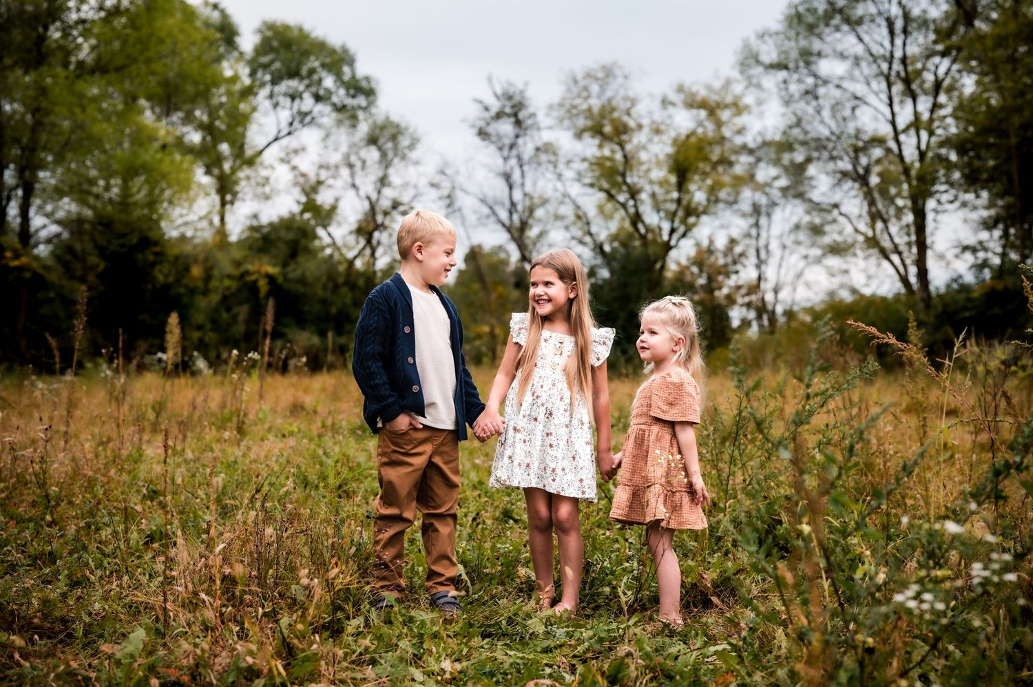 Three children smiling and laughing together in a sunlit meadow surrounded by tall grass and trees.