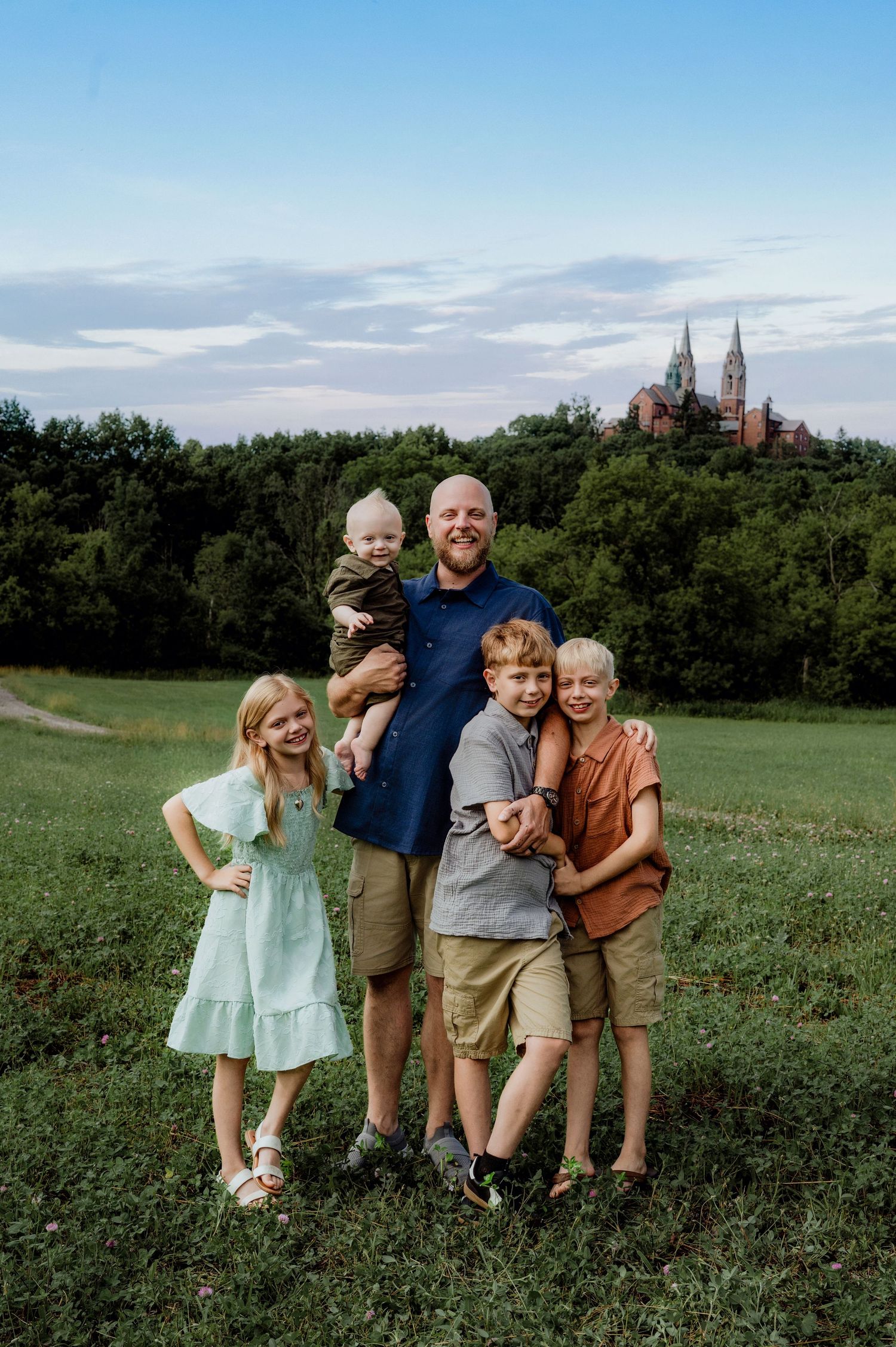 A father poses outdoors with four children on a green field, with a historic church visible in the background.