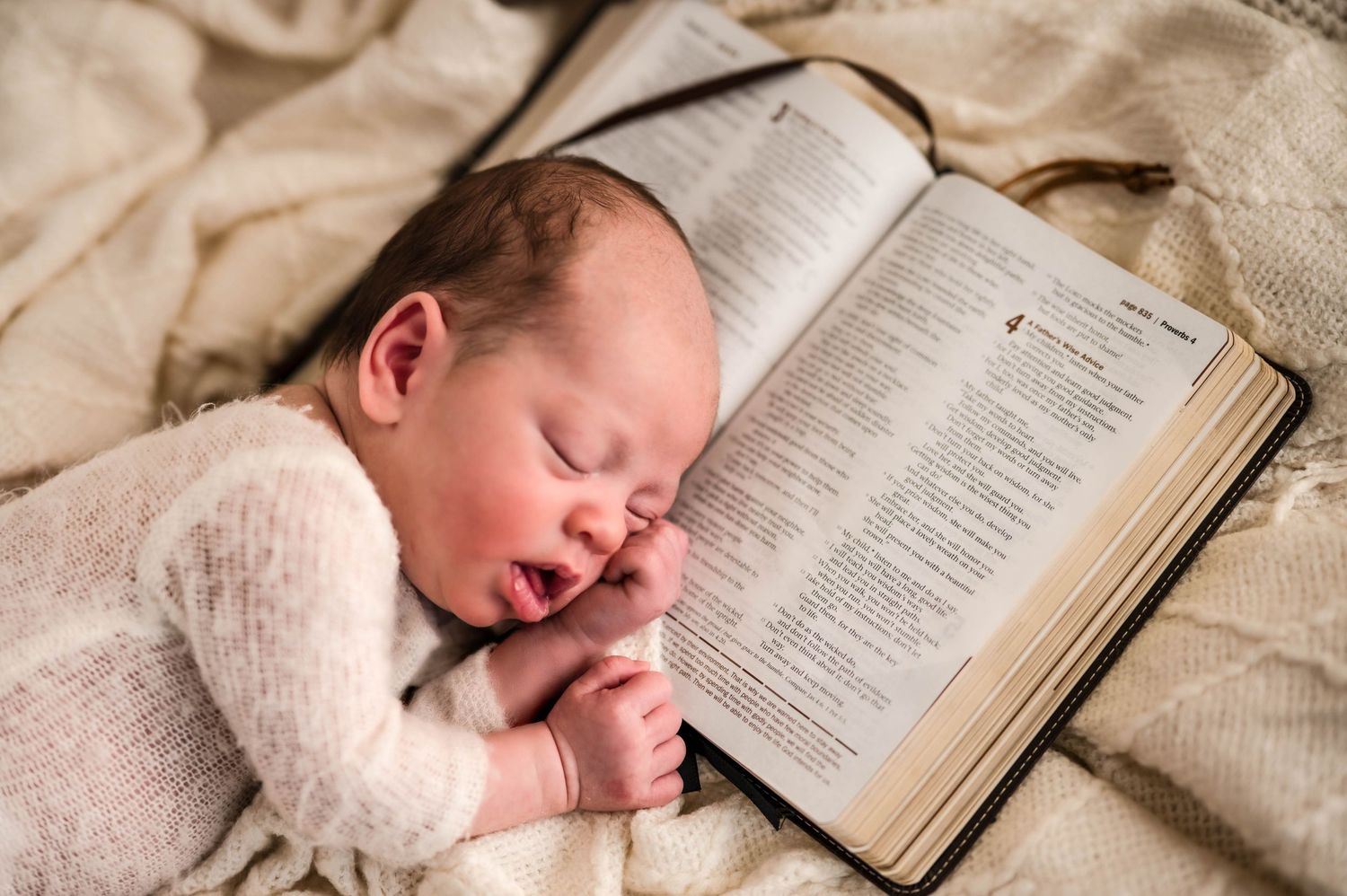 Sleeping newborn baby in a cream knit outfit resting its head on an open Bible on a soft beige blanket.