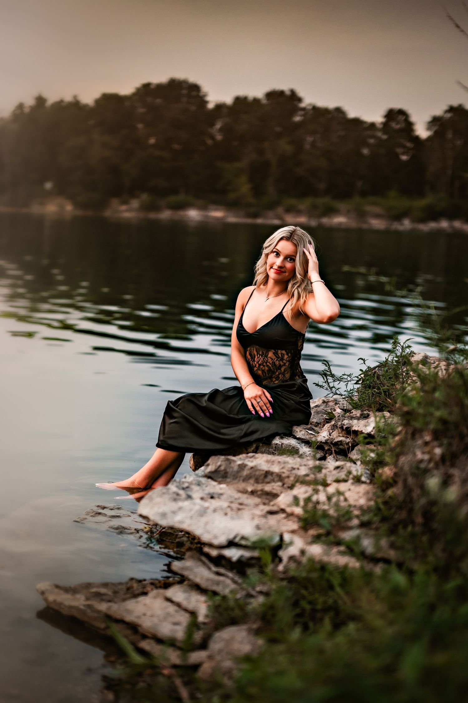 A woman in a black dress sits on rocky shoreline at sunset beside a serene lake with pine trees in background in Menomonee Falls, WI