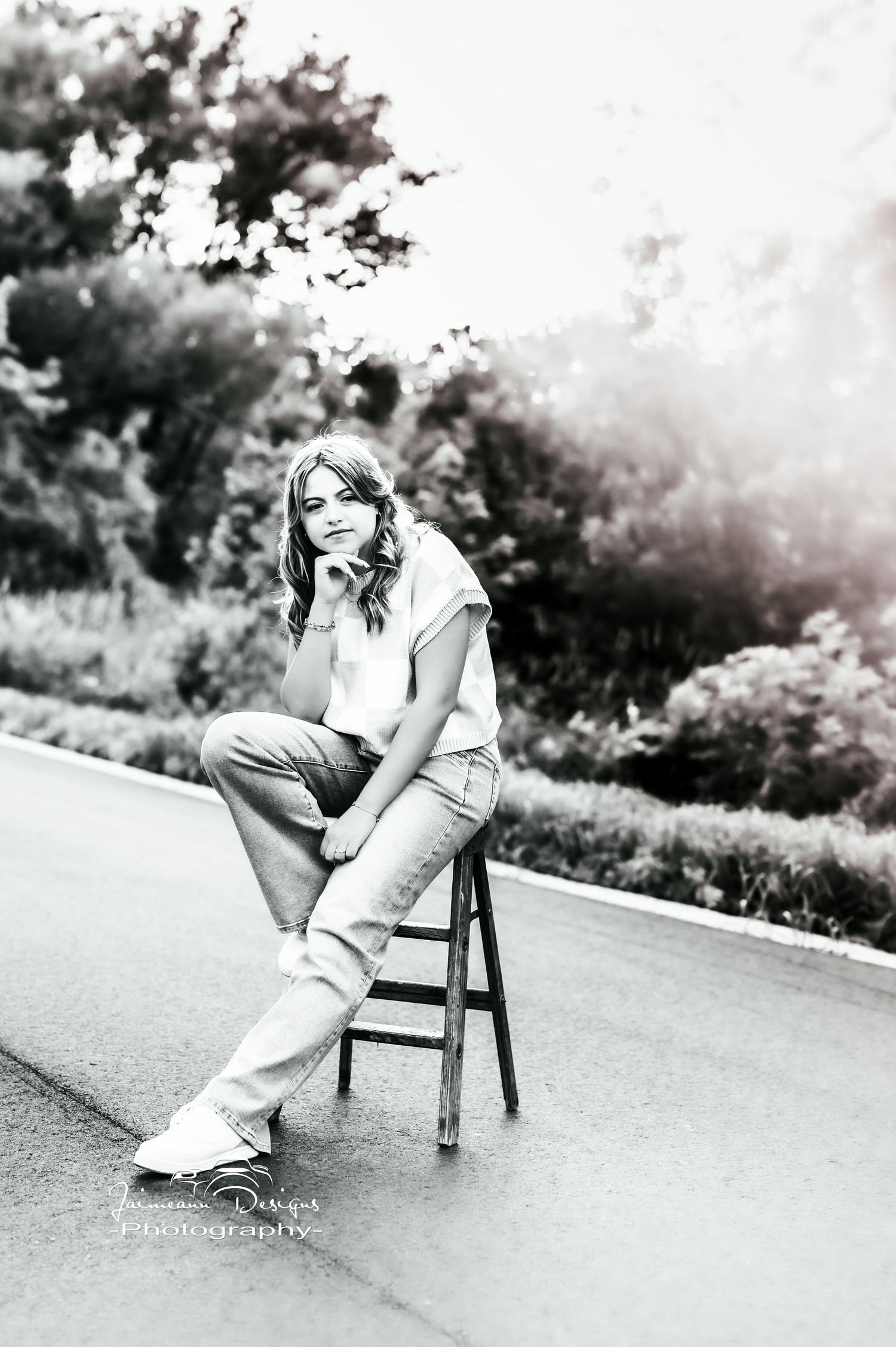 Black and white portrait of someone sitting casually on a wooden stool outdoors wearing jeans and a t-shirt.