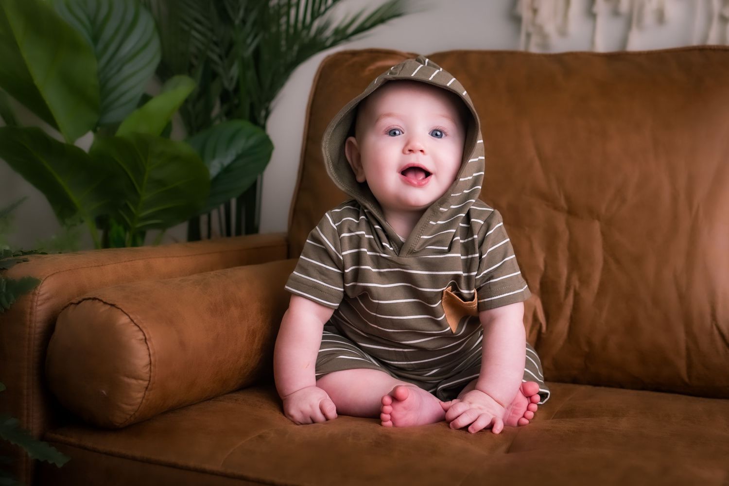 A cheerful baby in striped clothing sits on a brown leather sofa with plants in background.