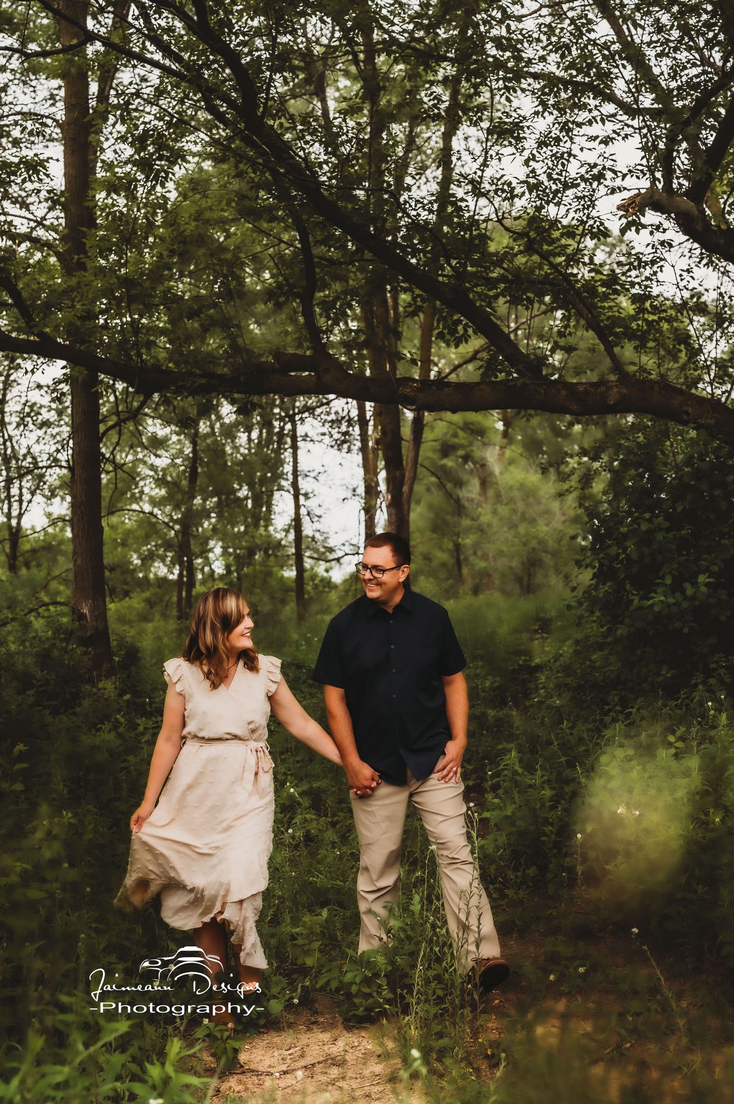 A couple holds hands while walking along a wooded path in casual attire.