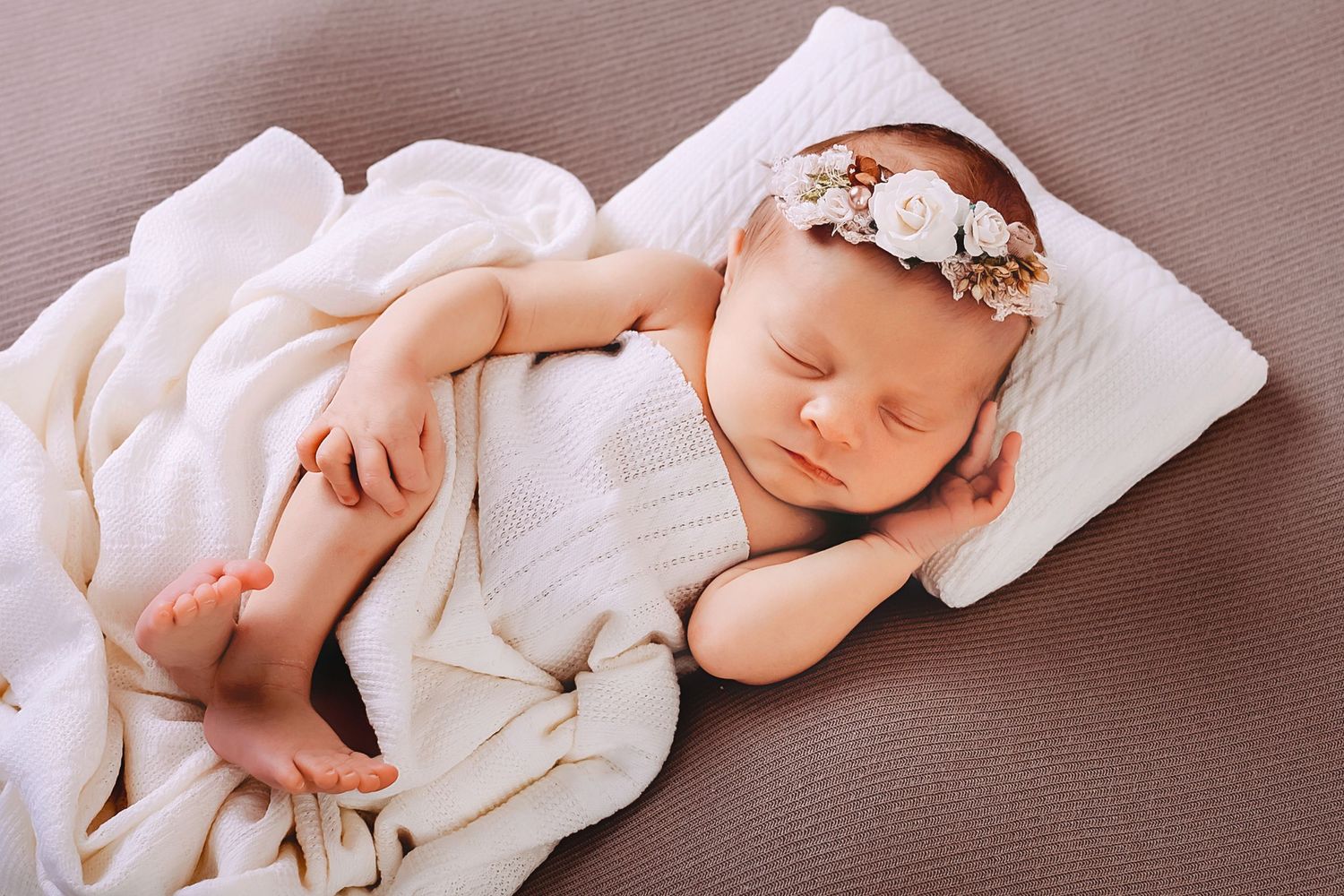 Peaceful newborn baby girl sleeping wrapped in white blanket with floral headband on soft brown background.