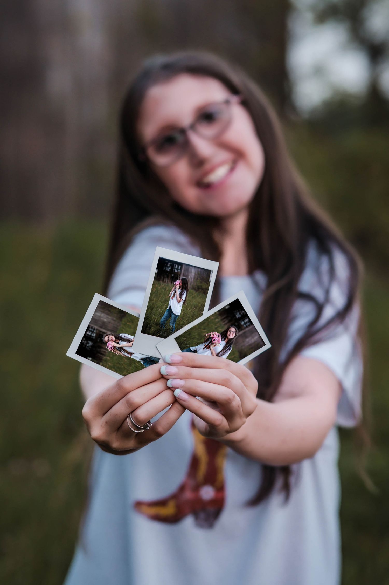 Smiling woman with glasses holds three Polaroid photos outdoors, showcasing candid snapshots in a blurred bokeh background.