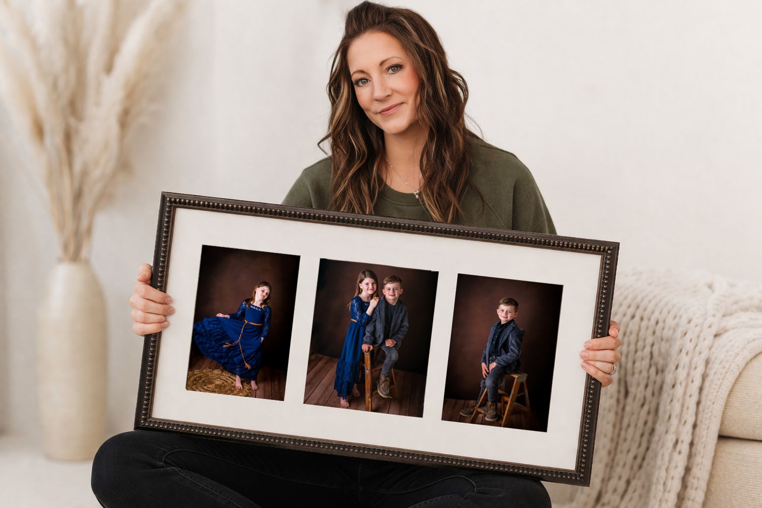 Woman smiling while holding a framed triptych portrait of children in blue outfits against a warm studio background.