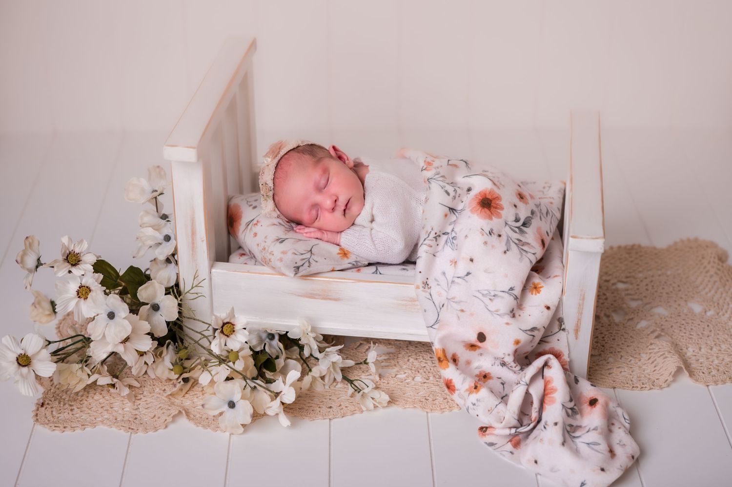 Sleeping newborn baby in white swaddle blanket surrounded by delicate flowers and lace decorations on neutral backdrop.