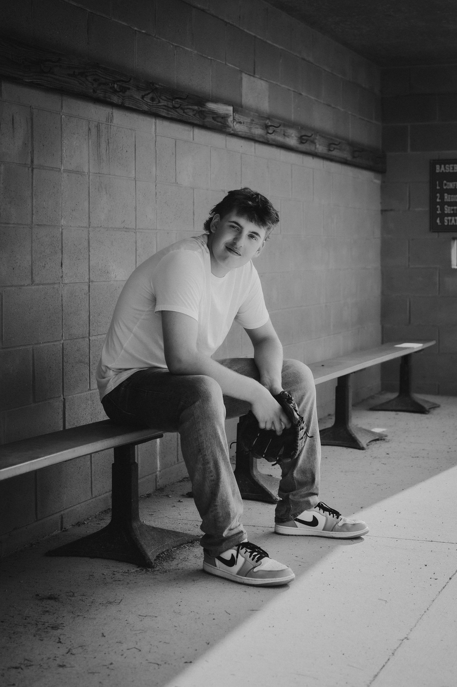 Young man in white t-shirt and Nike sneakers sits on a bench in a dugout, holding a camera, black and white photo.