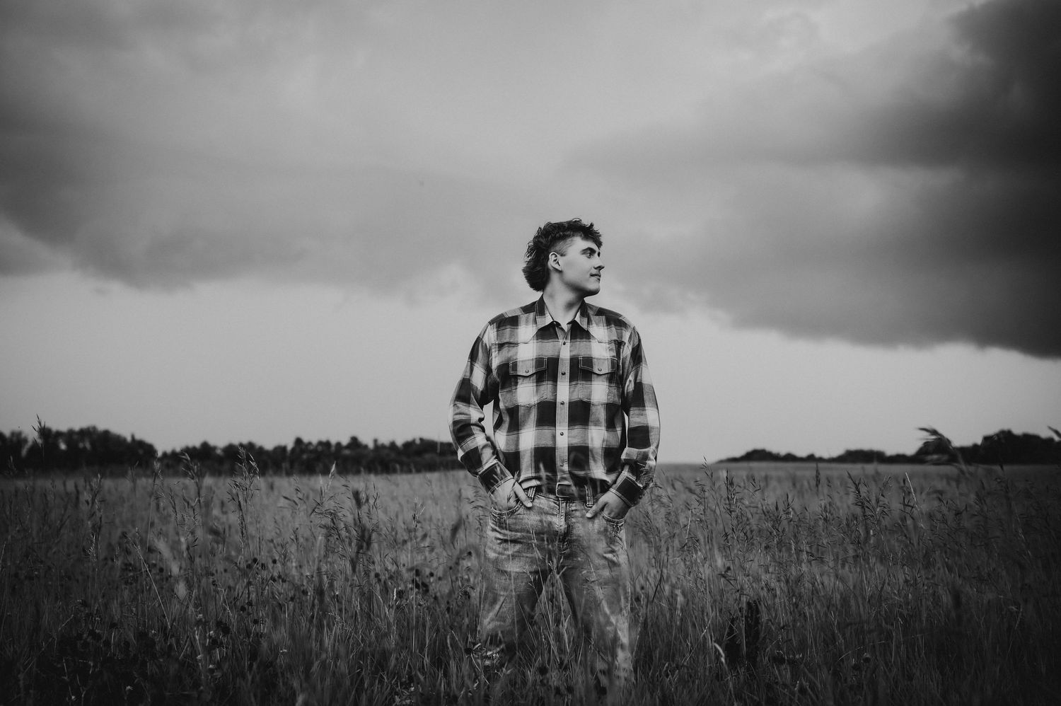 Black and white photo of a young man standing in a field wearing a plaid shirt under dramatic stormy skies.