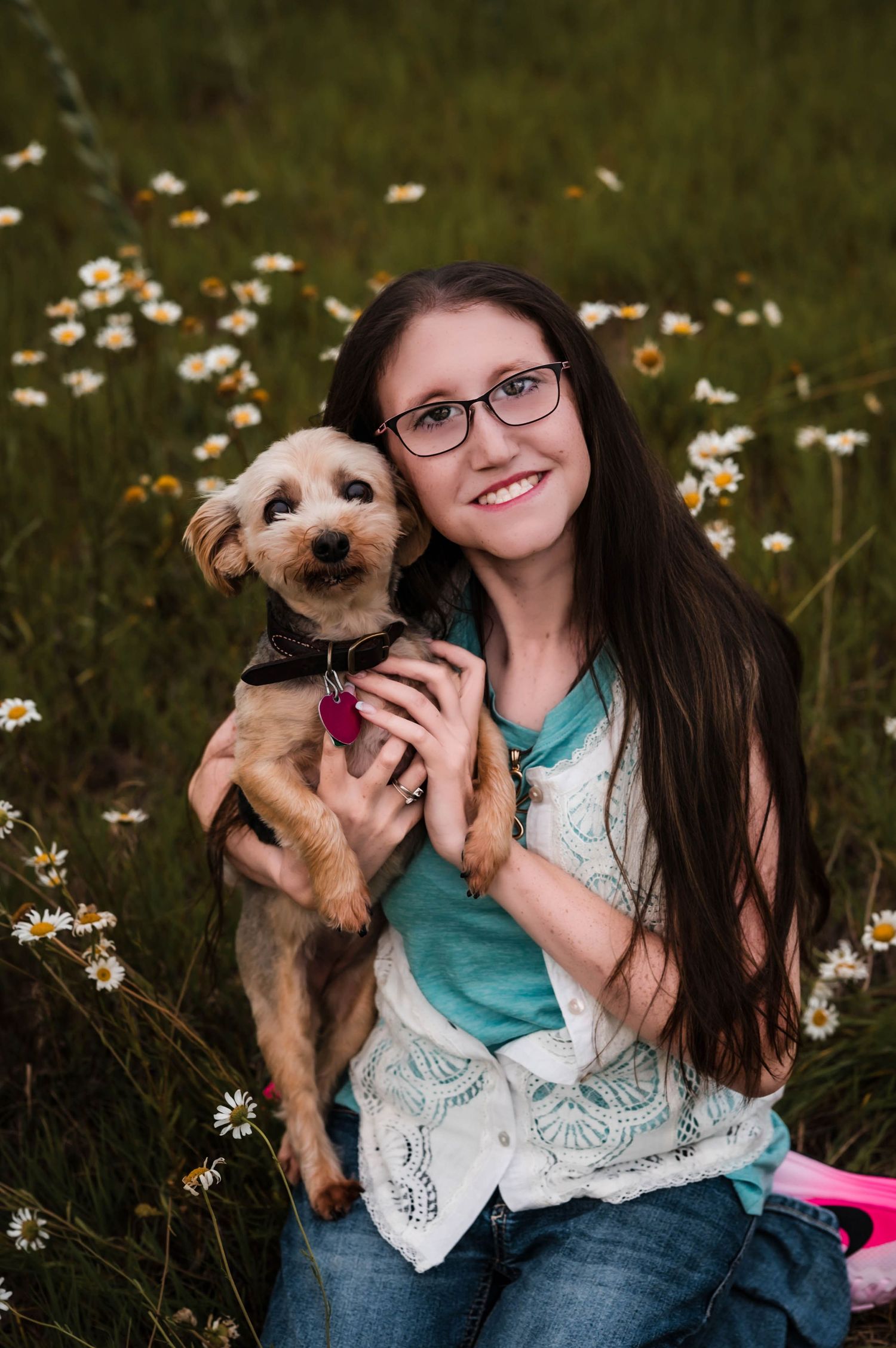 Young woman with long dark hair and glasses smiles holding a small terrier dog in a daisy field.