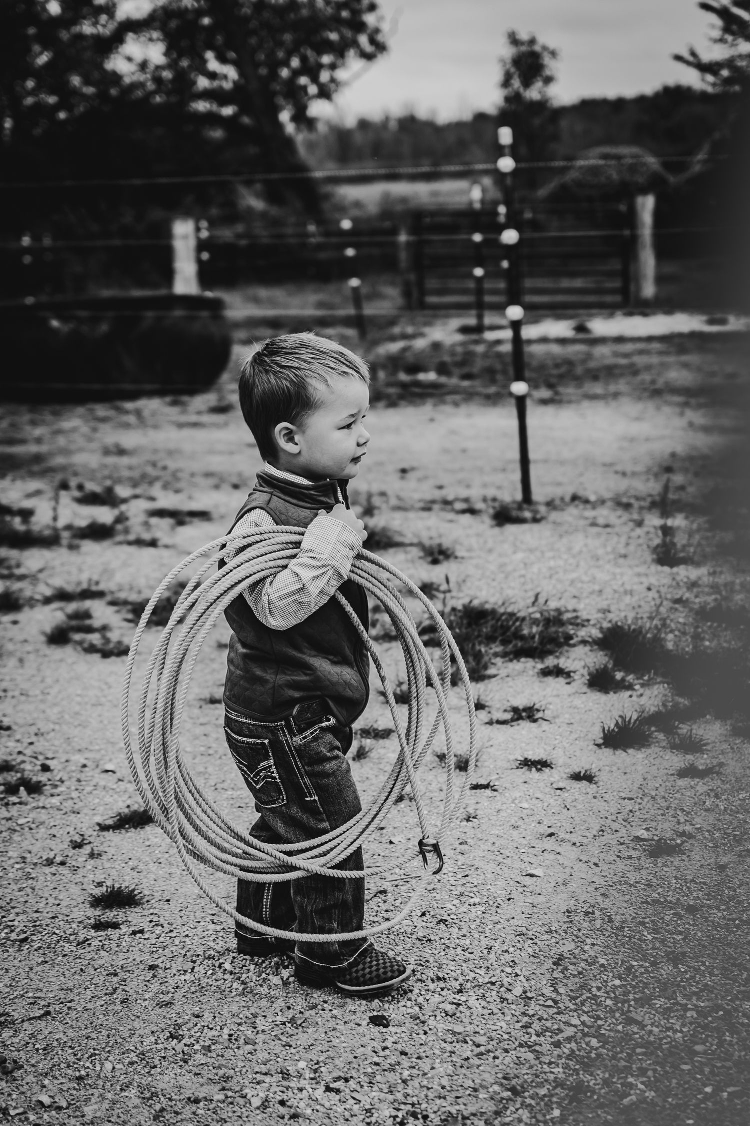Black and white photo of a small child holding a lasso rope on rustic farmland.