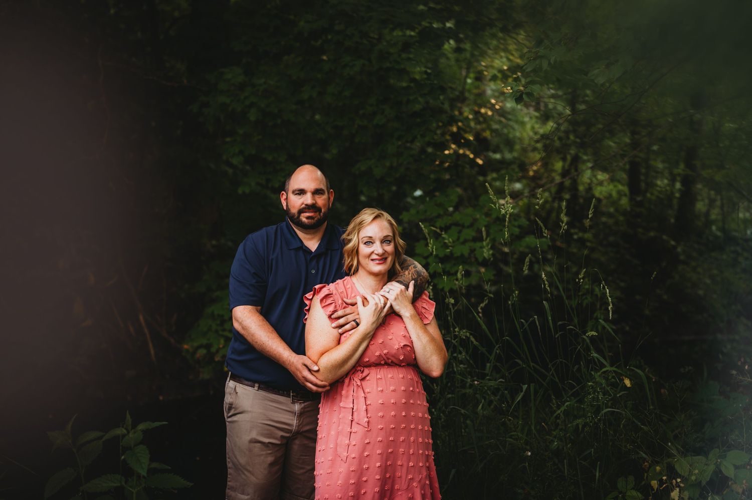 Couple embraces in outdoor portrait against green forest background.