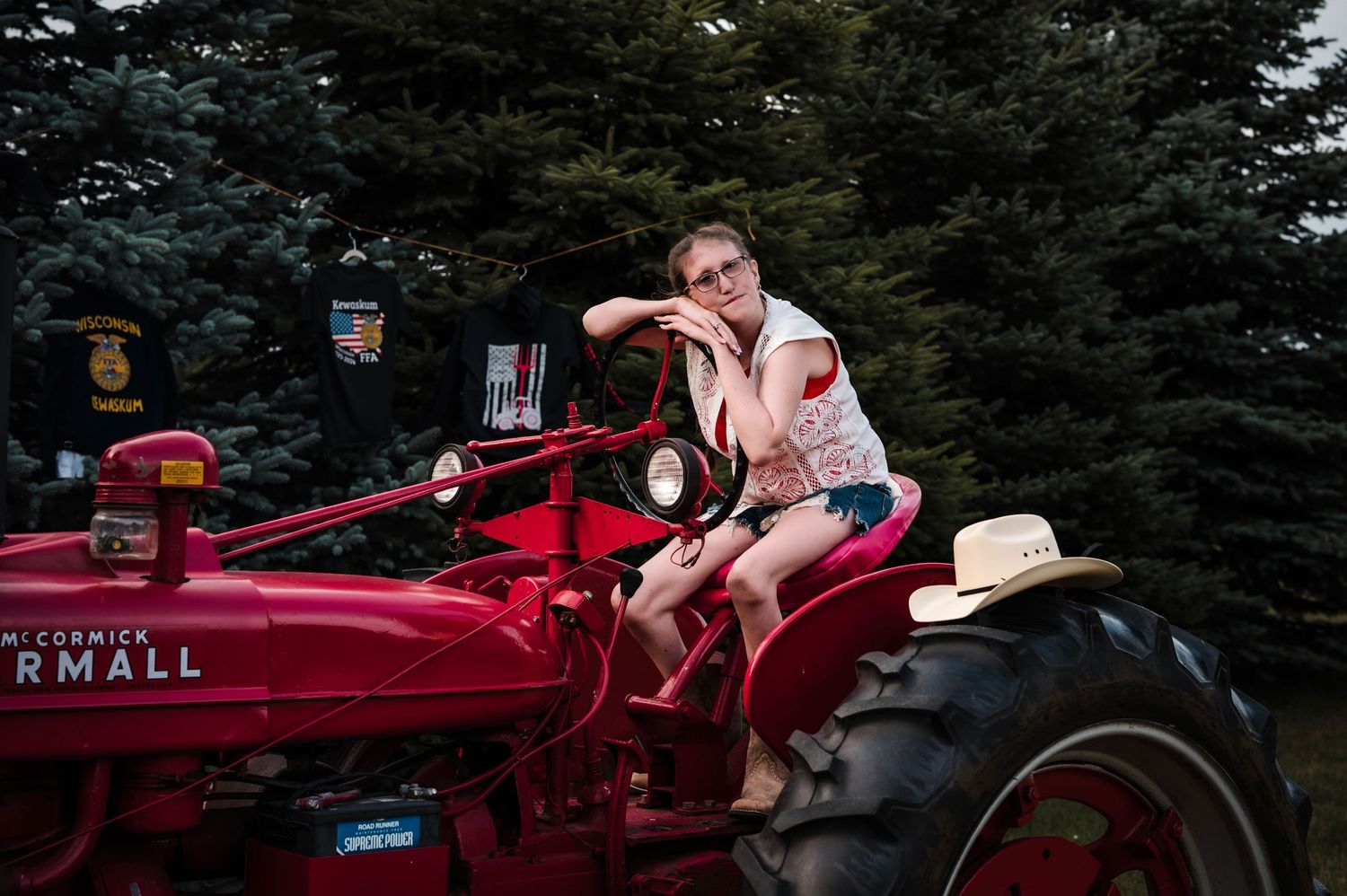 Young girl sitting on a red Farmall McCormick tractor outdoors near evergreen trees wearing glasses and denim shorts.
