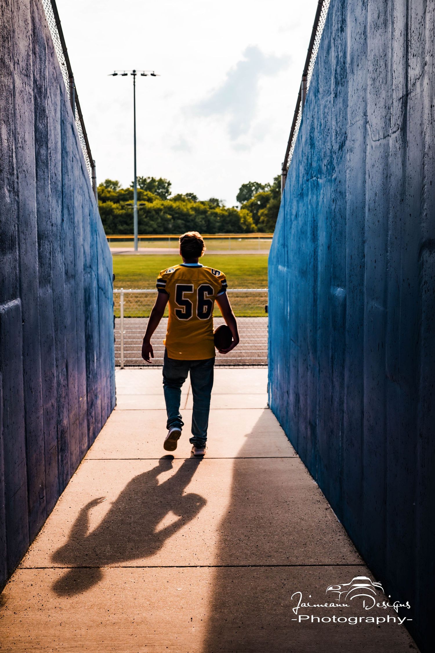 Football player #56 in yellow jersey walks through stadium tunnel carrying helmet, casting dramatic shadow on concrete floor.