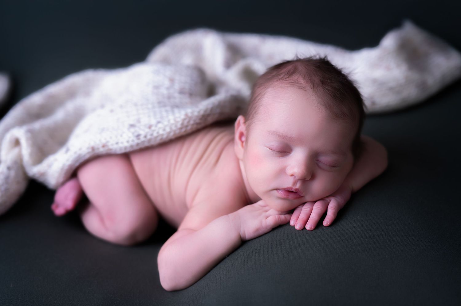 Peaceful newborn baby sleeping on stomach with white knit blanket on dark background.