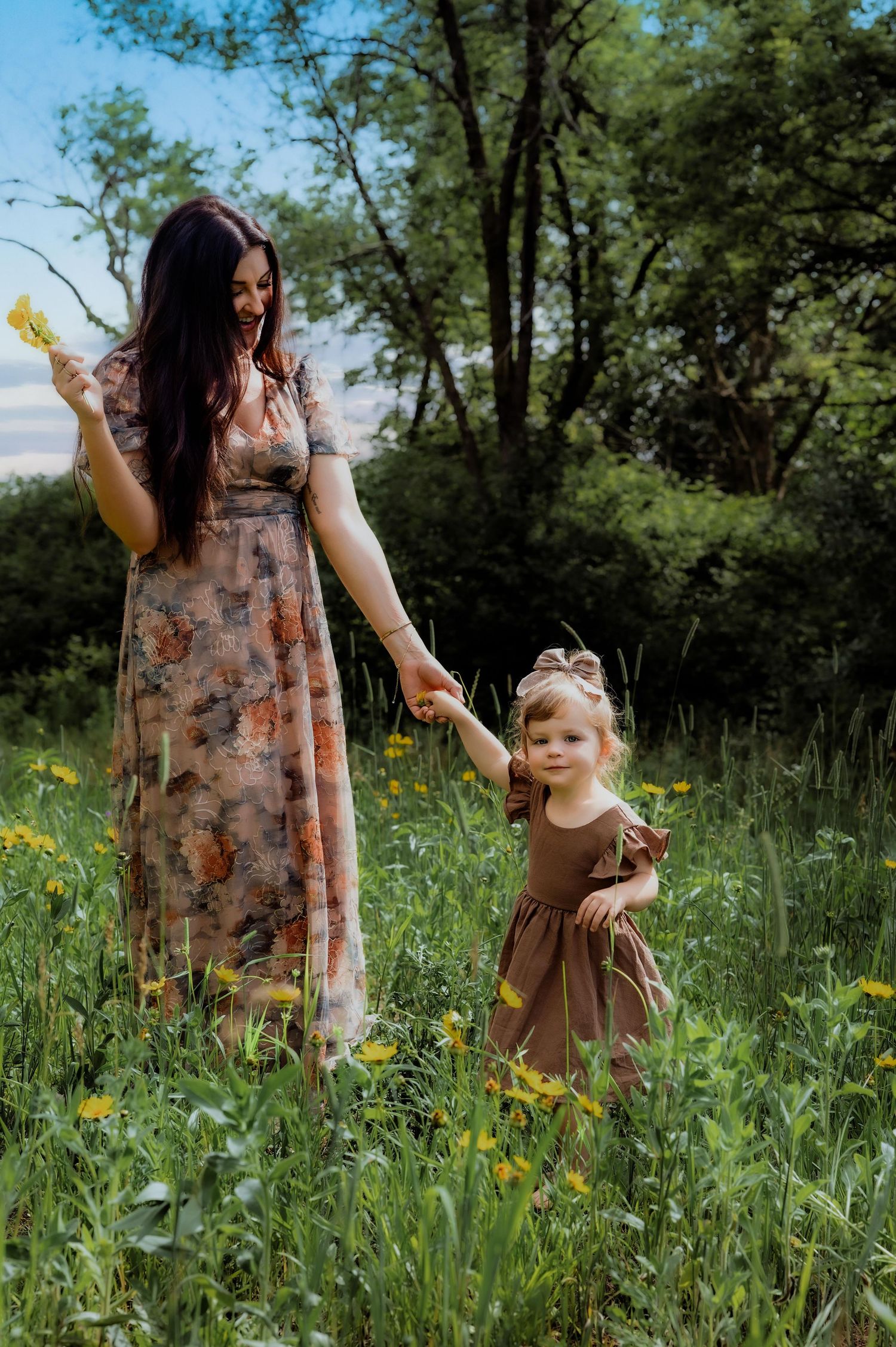 Mother in floral dress holds hands with toddler daughter in brown dress walking through wildflower meadow.