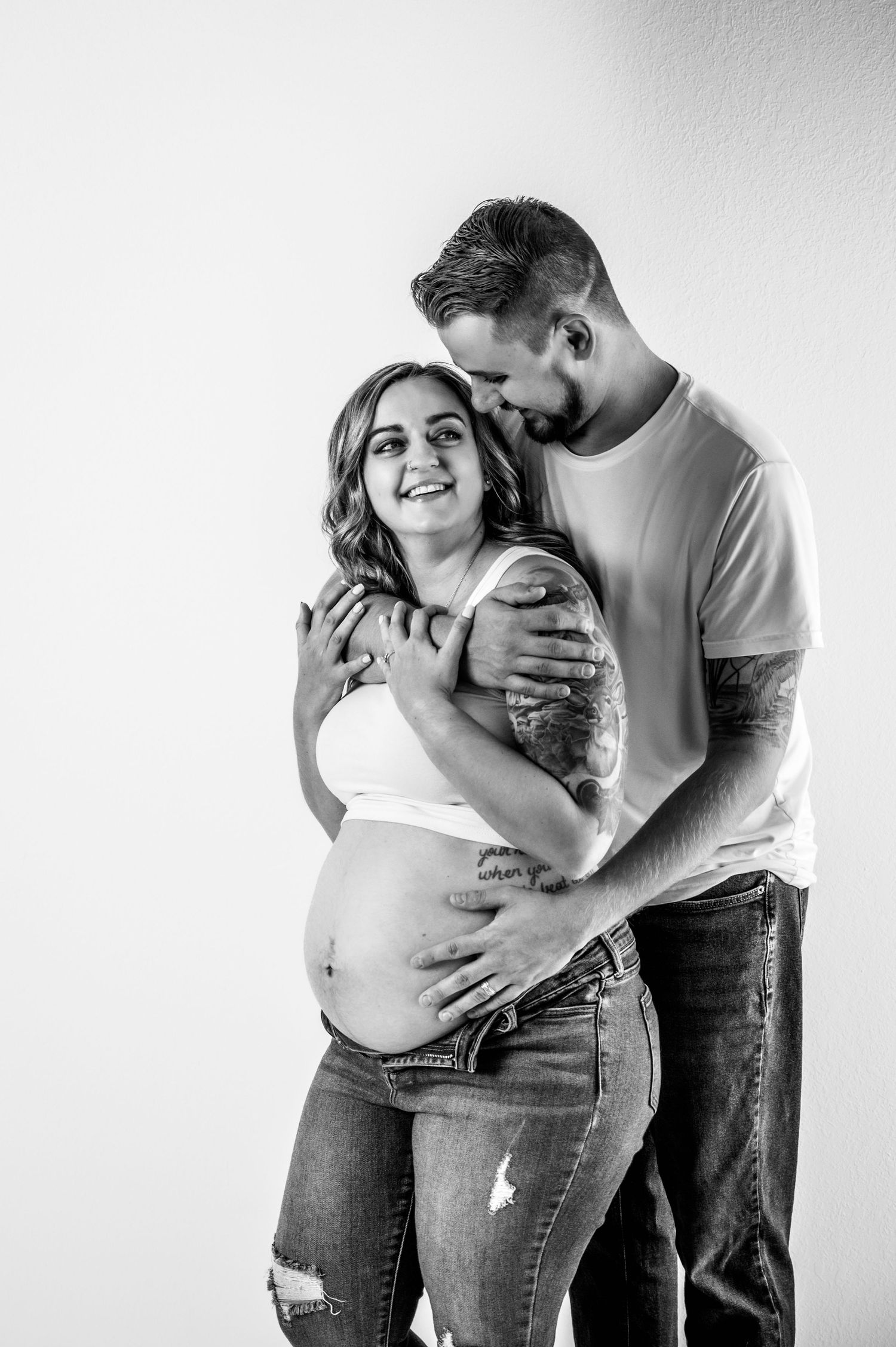 Black and white maternity photos show an intimate moment between expecting parents in casual jeans and t-shirts.