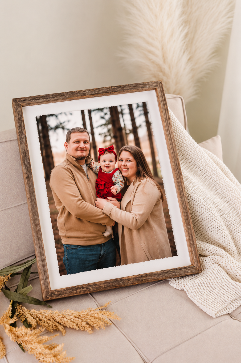 A rustic wood-framed family portrait of a couple holding a baby in red, displayed on a light surface with pampas grass decor.
