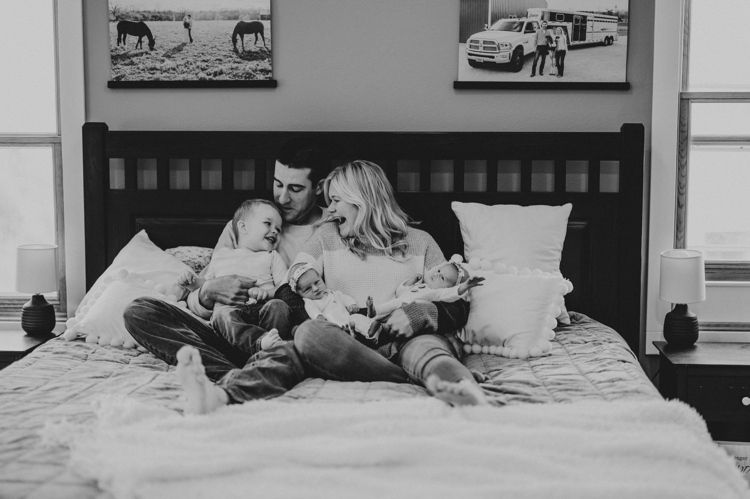 Black and white photo of a couple sitting on a bed with their newborn baby in a cozy bedroom setting.