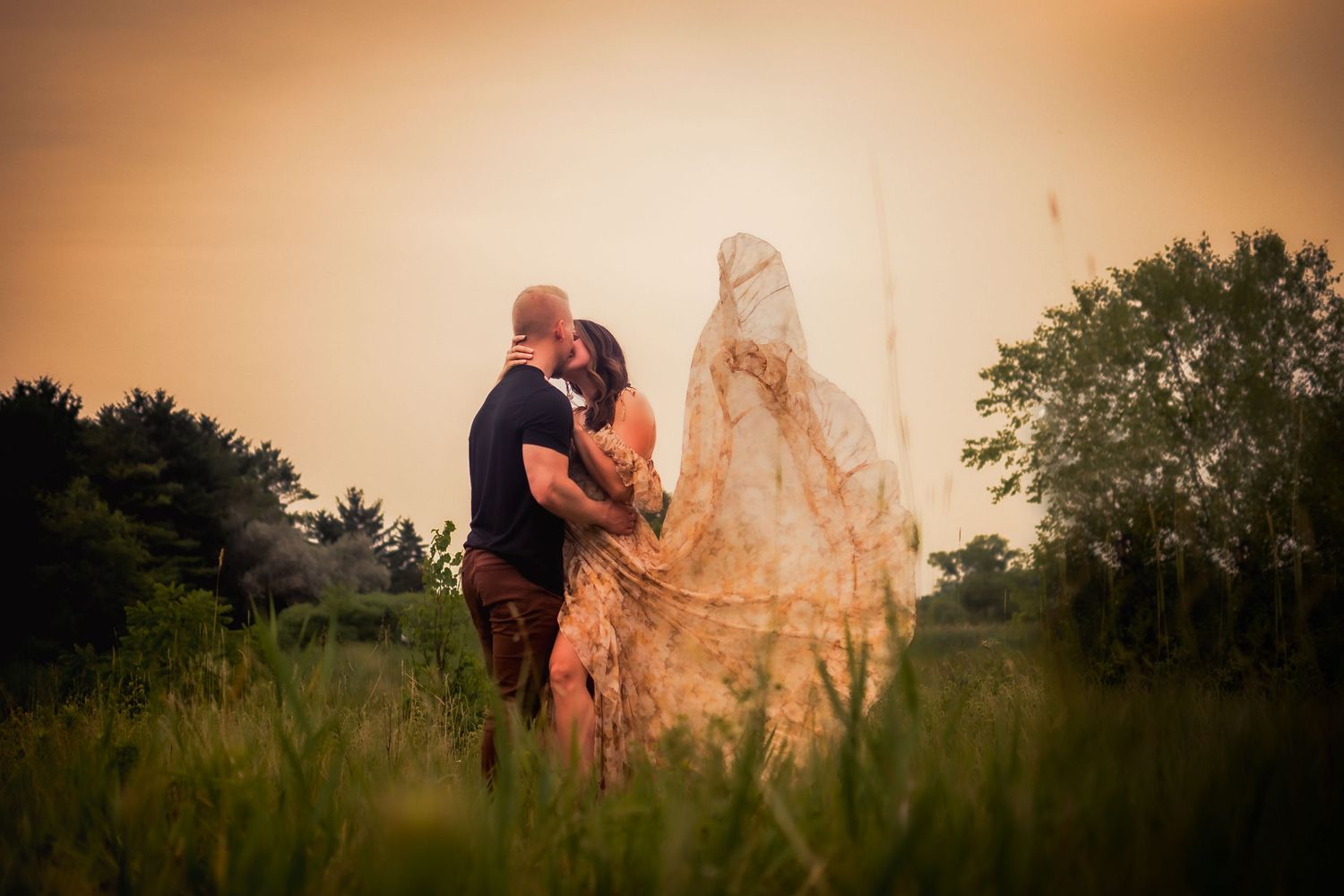 Couple embracing in golden field with flowing dress during sunset.