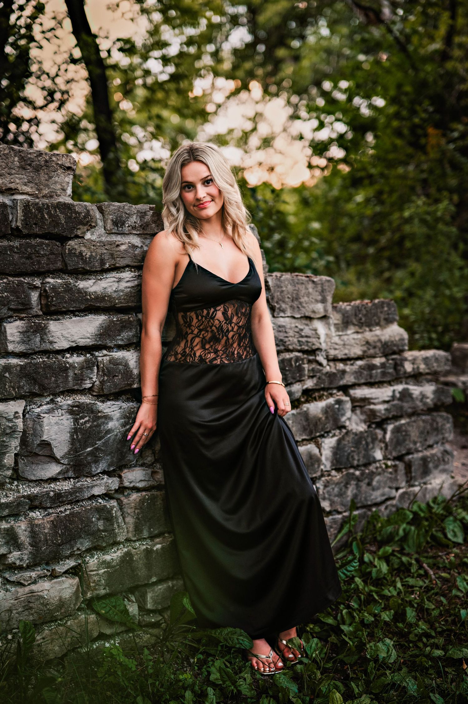 A woman in a black dress poses against a stone wall at sunset in an outdoor portrait session in Quarry Park in Menomonee Fall, WI