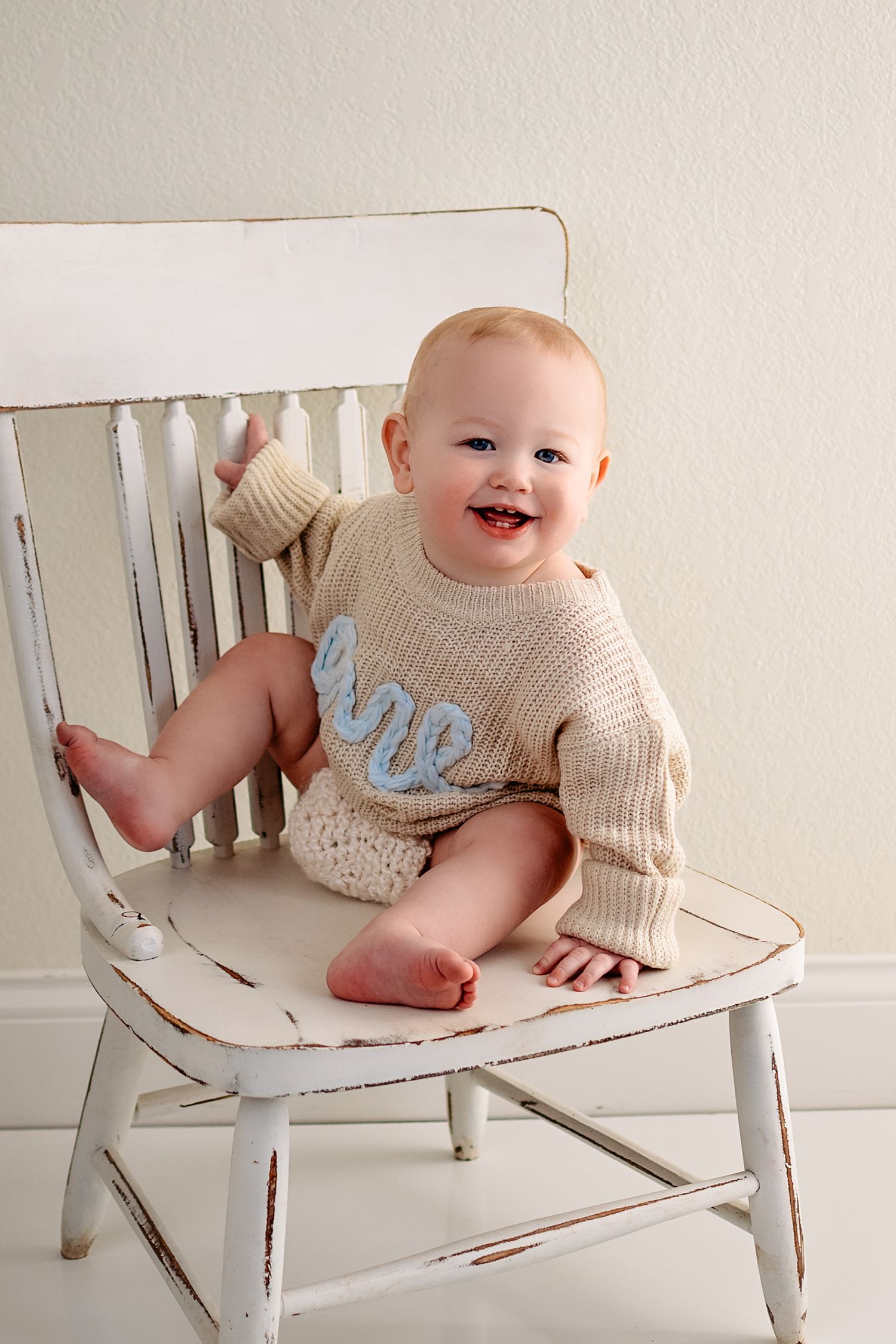 A baby wearing a knit sweater sits and smiles on a distressed white wooden bench in a progression of playful poses near Campbellsport, WI