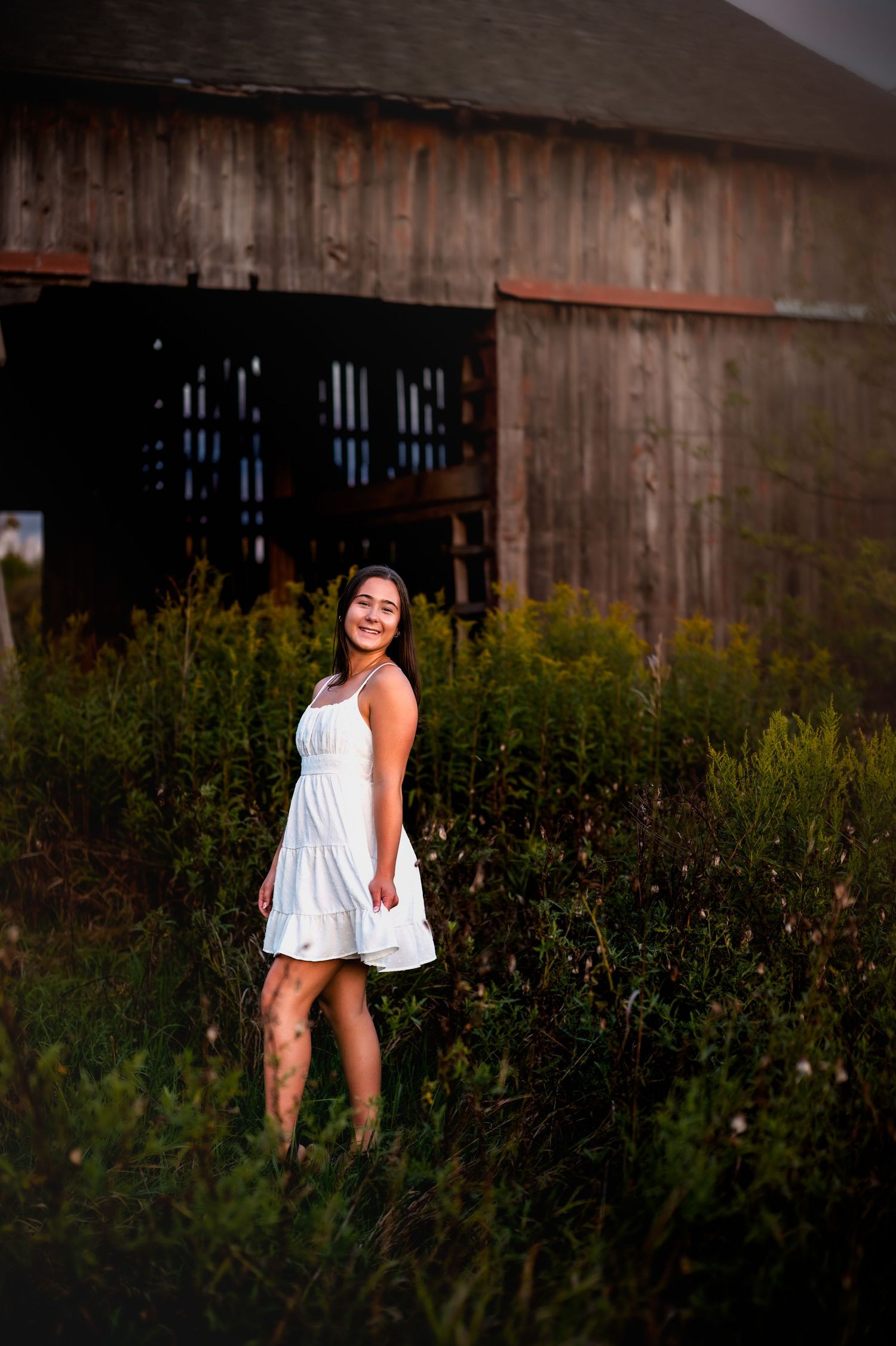 A woman in a white dress poses playfully in a field of wildflowers against a rustic barn backdrop near Boltonville, WI