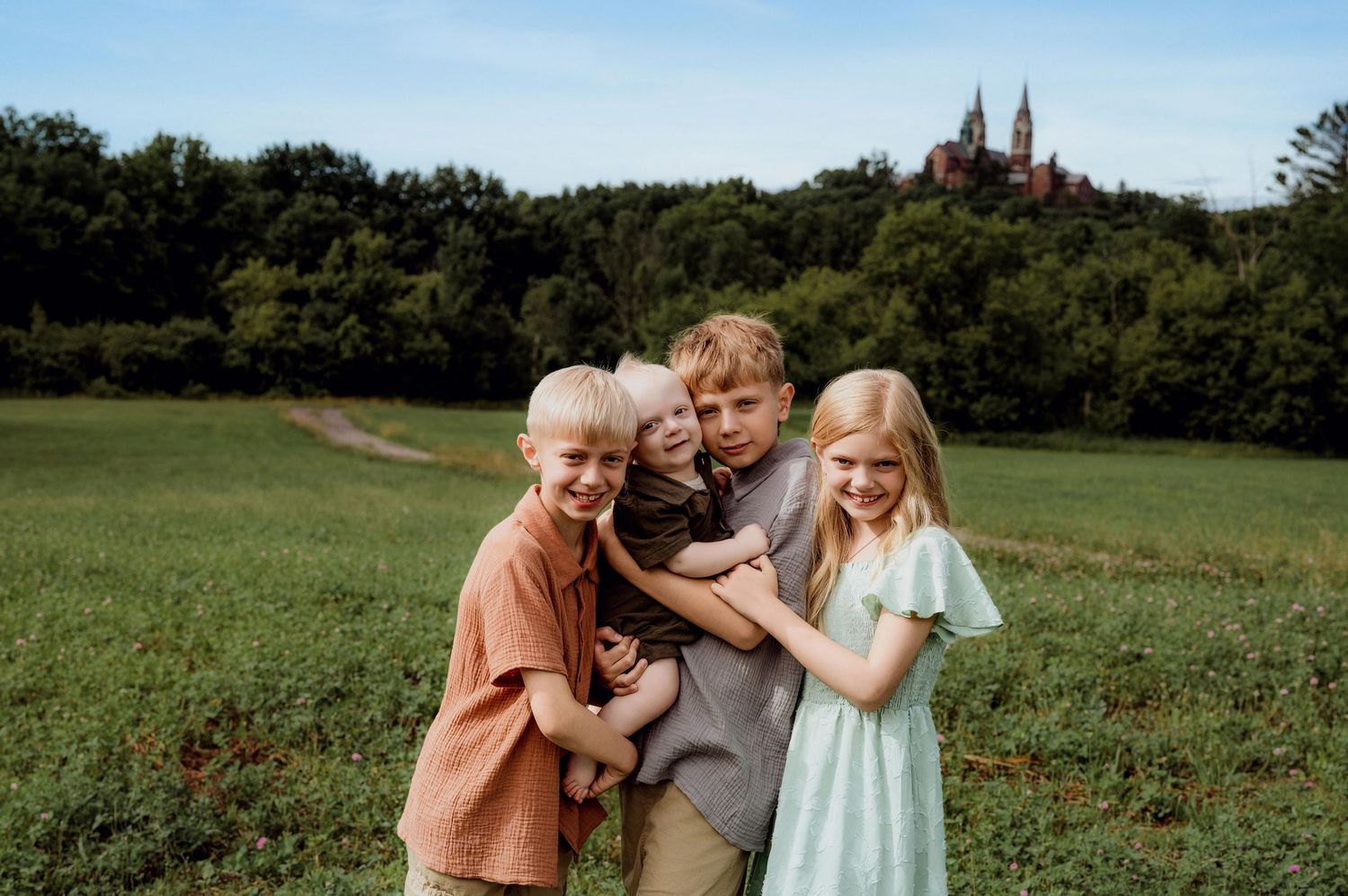 Four children smiling and hugging in a green field with a historic church on a hill in the background.
