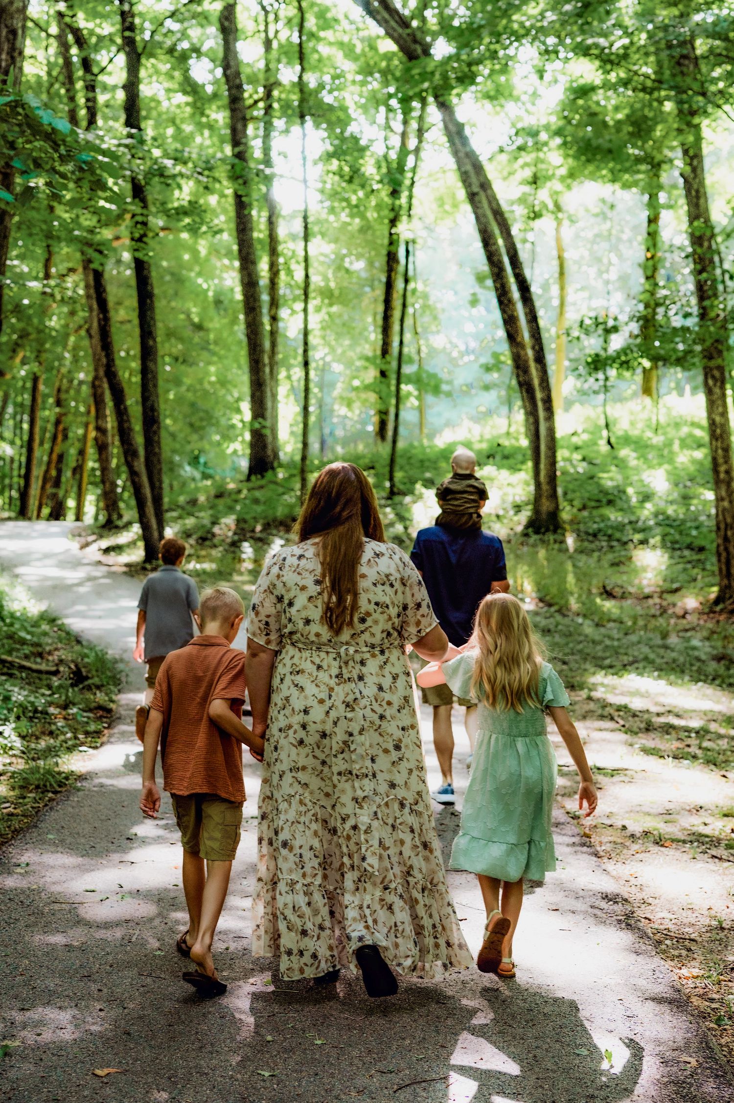 Family walking together on a shaded forest path surrounded by lush green trees on a sunny day.