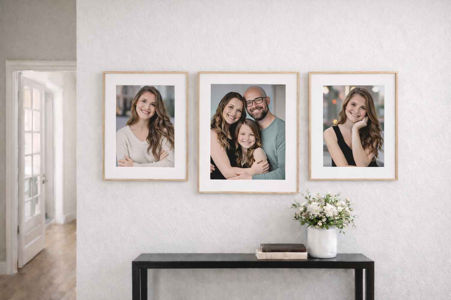 Three framed family portrait photos displayed on a white wall above a black console table with white flowers.