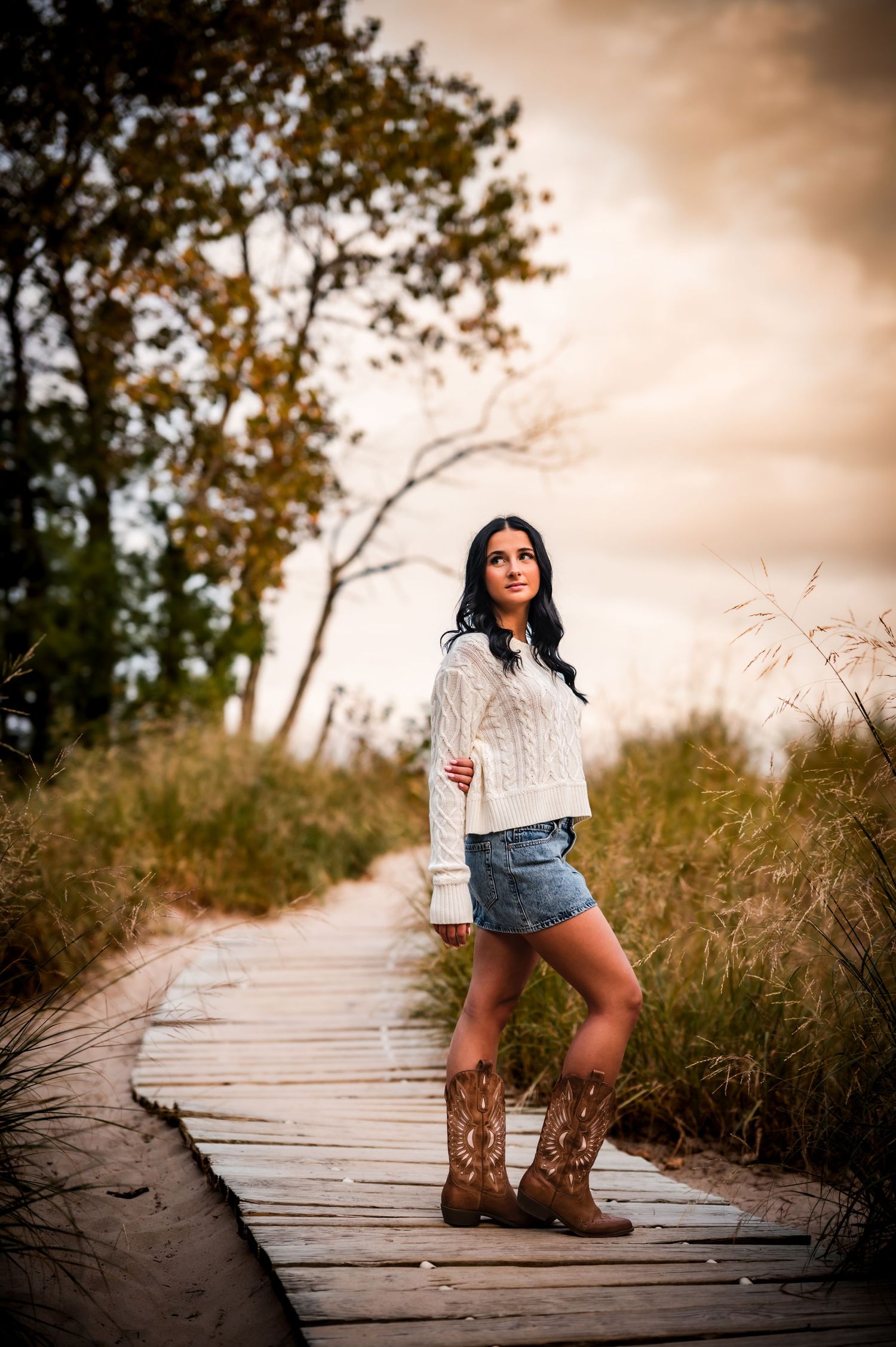 A person in a white sweater and denim shorts poses on a wooden boardwalk wearing brown cowboy boots at sunset.