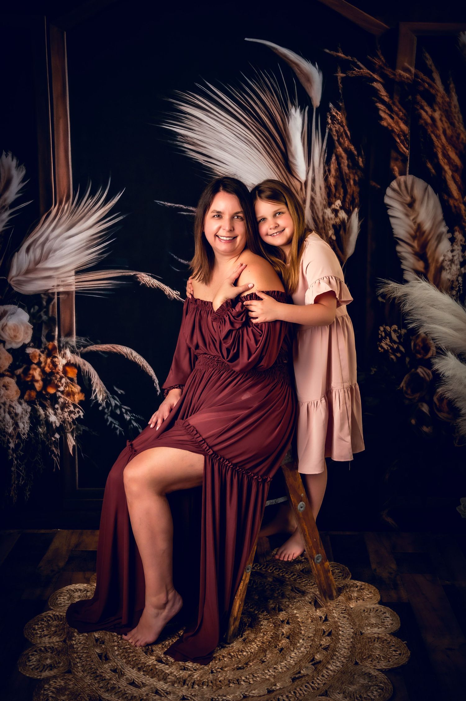 Elegant maternity photoshoot featuring burgundy gown and dried palm decorations against dark backdrop.