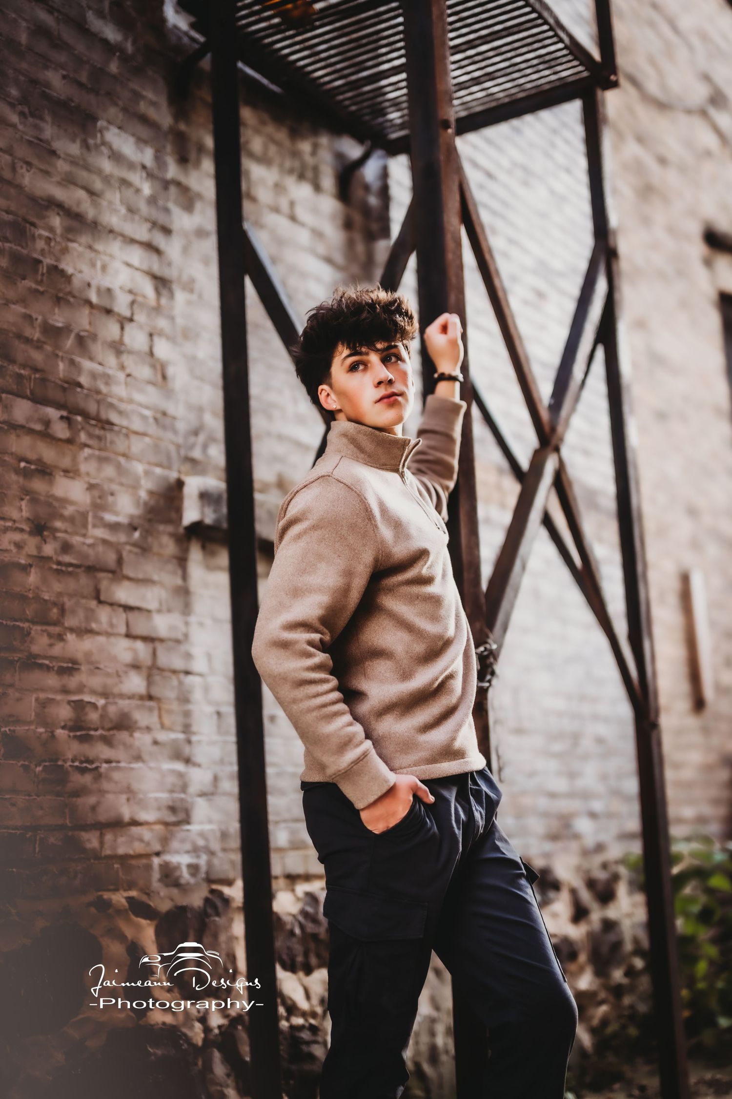 Stylish casual portrait against industrial brick wall backdrop featuring tan sweater and dark pants.
