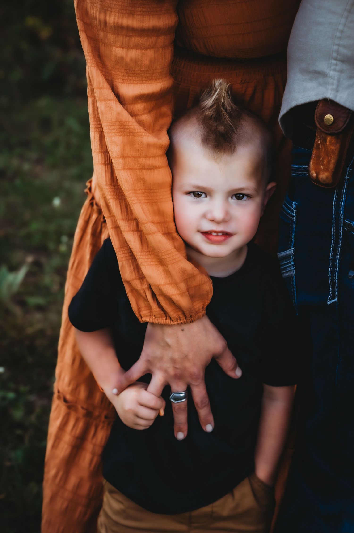 A small child peeks out from behind someone wearing orange pants while holding onto their hand with a bright smile.
