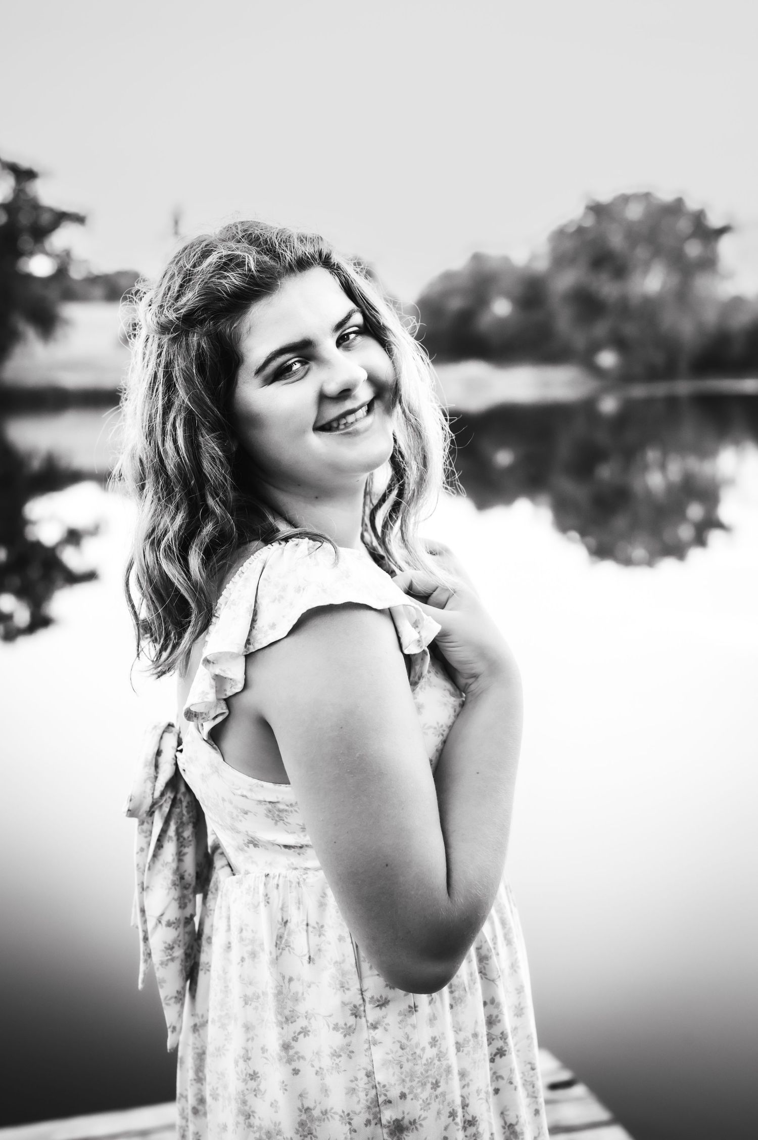 Black and white portrait of someone in a floral dress smiling by a lake at sunset.