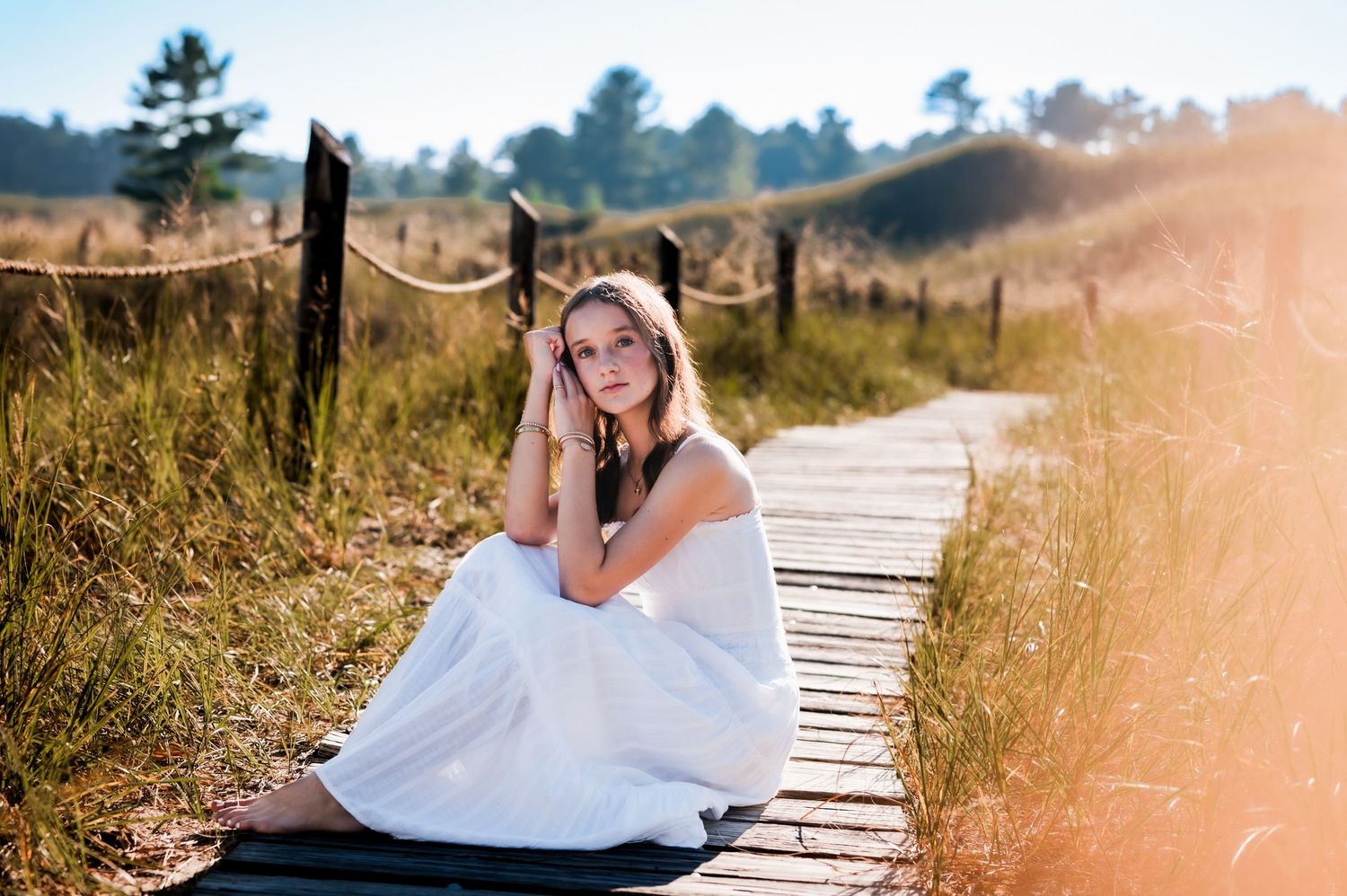 A white dress adorns a figure seated on a wooden boardwalk path through tall grass at golden hour in Delafield, WI