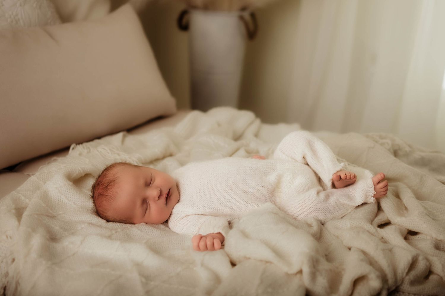 Sleeping newborn baby in a white outfit lying on a soft cream knit blanket in a bright, cozy room.