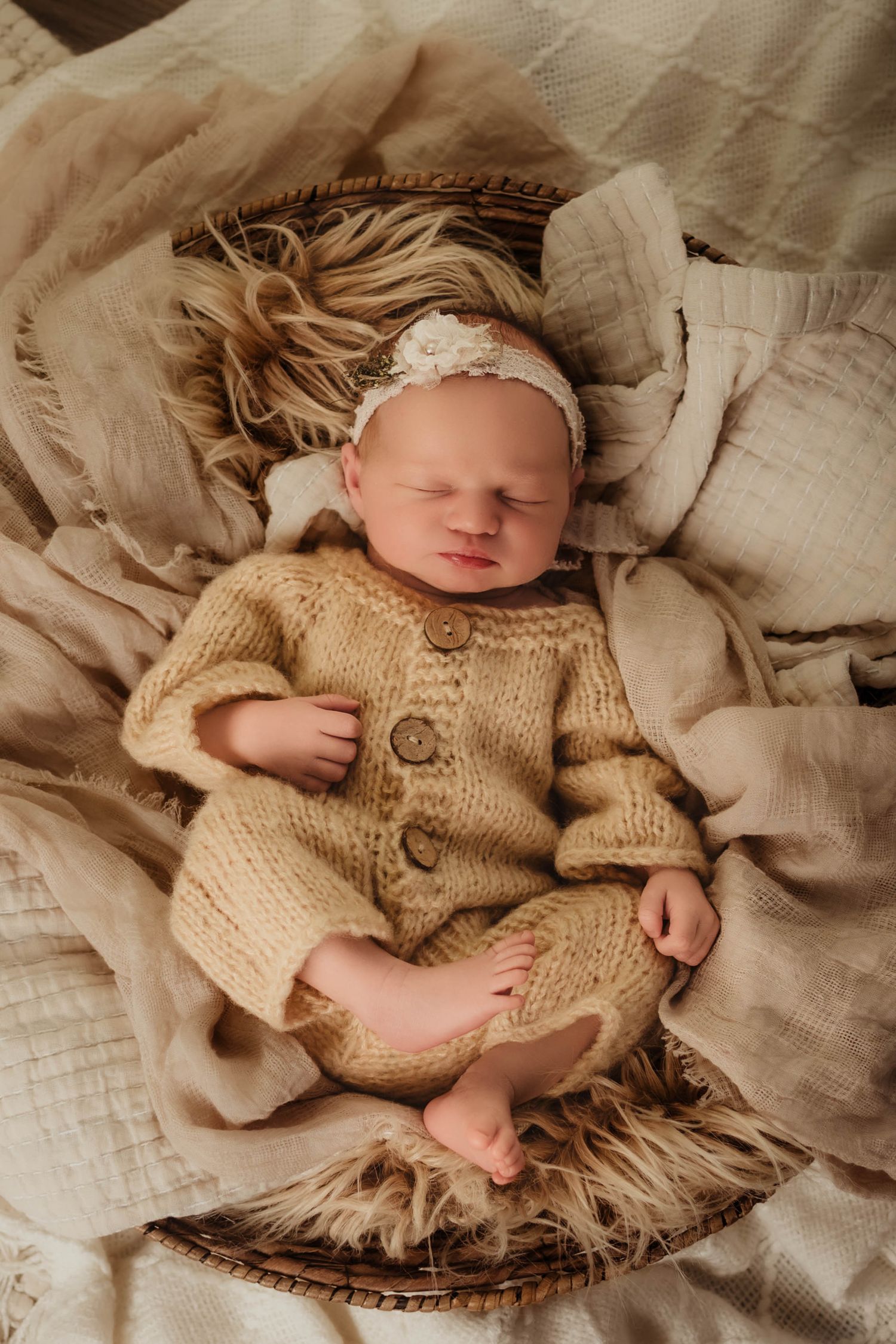 Sleeping newborn baby in a knit tan outfit and floral headband resting in a woven basket with fluffy fur and cream blankets.