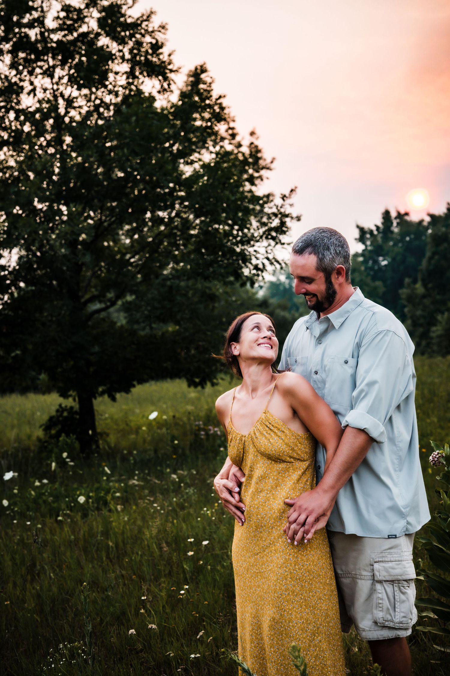 Couple embracing in a wildflower meadow at sunset, woman in yellow dress smiling up at man in light blue shirt.
