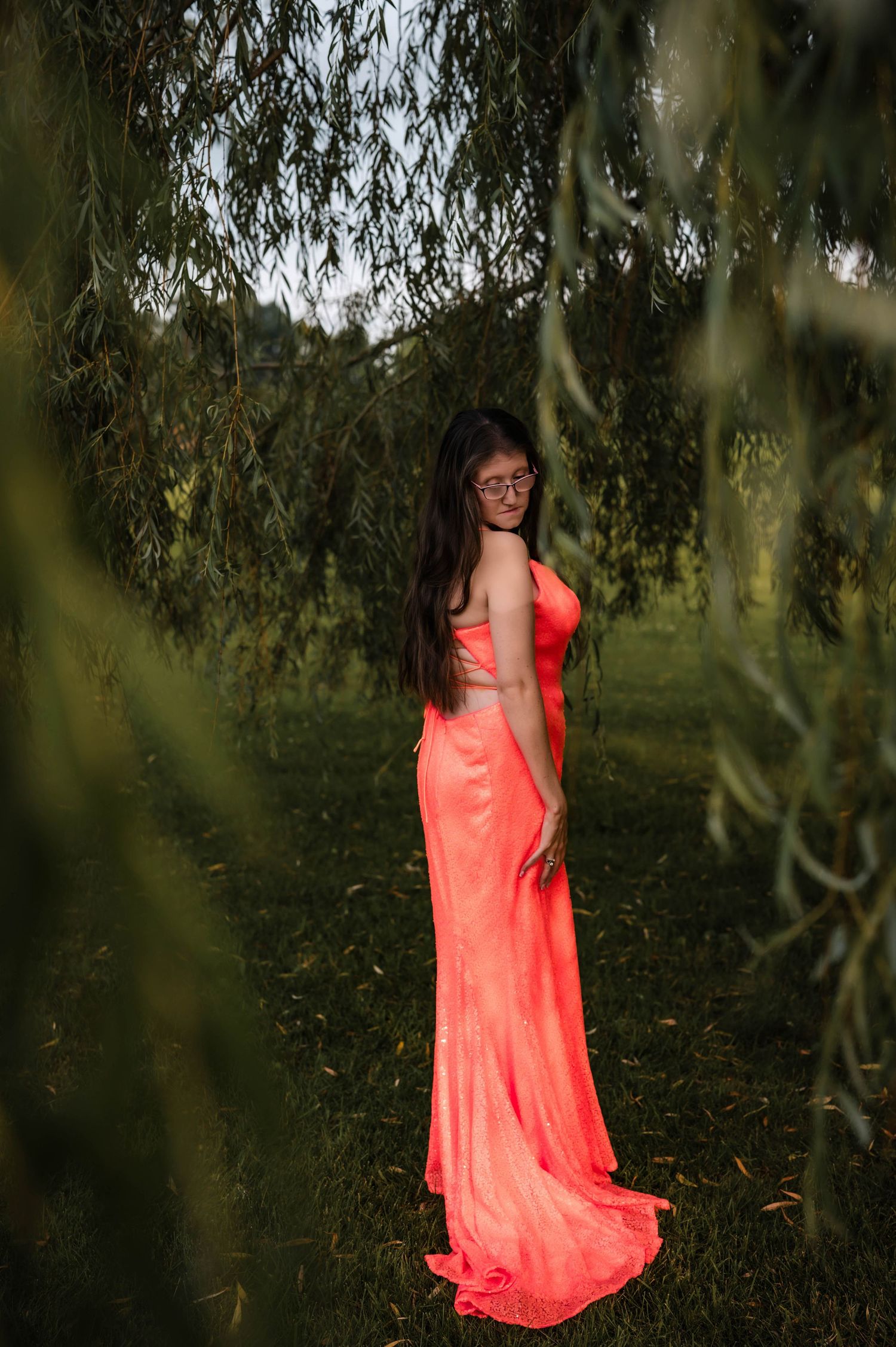 A woman in a flowing coral strapless gown stands gracefully beneath weeping willow branches in a lush green garden.