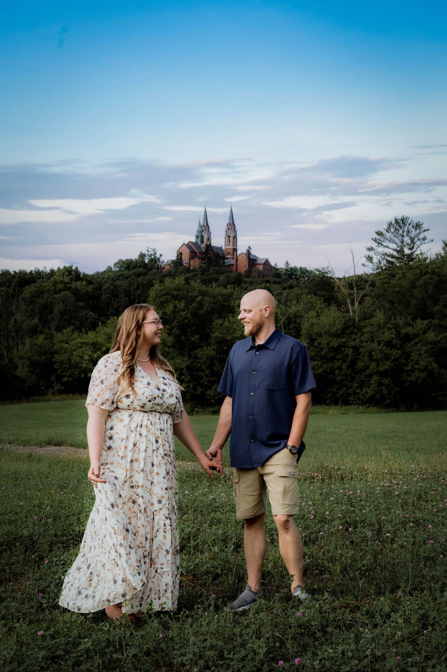 Couple holding hands in a green field with a historic church on a hill and blue sky backdrop.