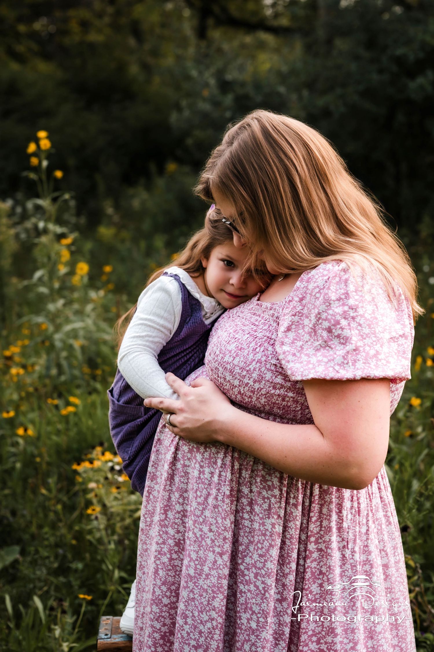 Pregnant woman in floral dress tenderly holds toddler in purple dress among wildflowers and yellow blooms outdoors.