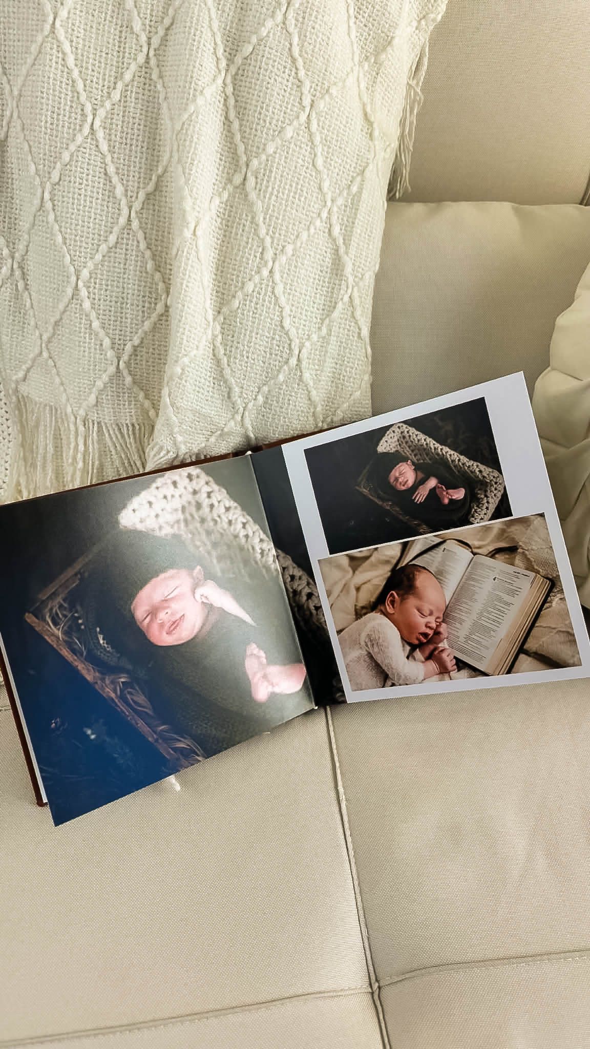 Open newborn photo album on a cream sofa showing sleeping baby portraits, one with an open book, beside a cream knit throw blanket.