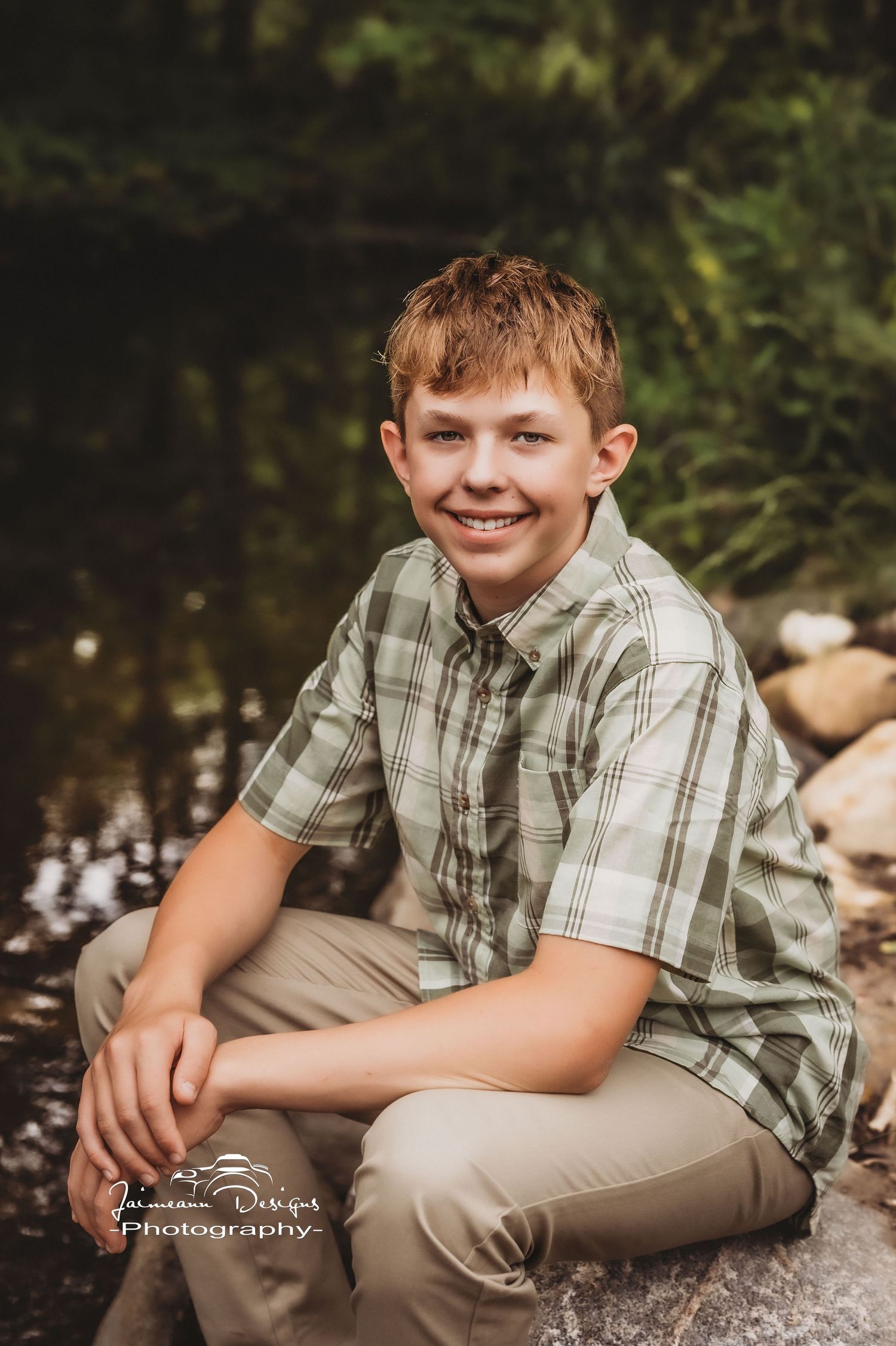 A casual portrait of someone in a plaid shirt sitting on rocks by a stream.