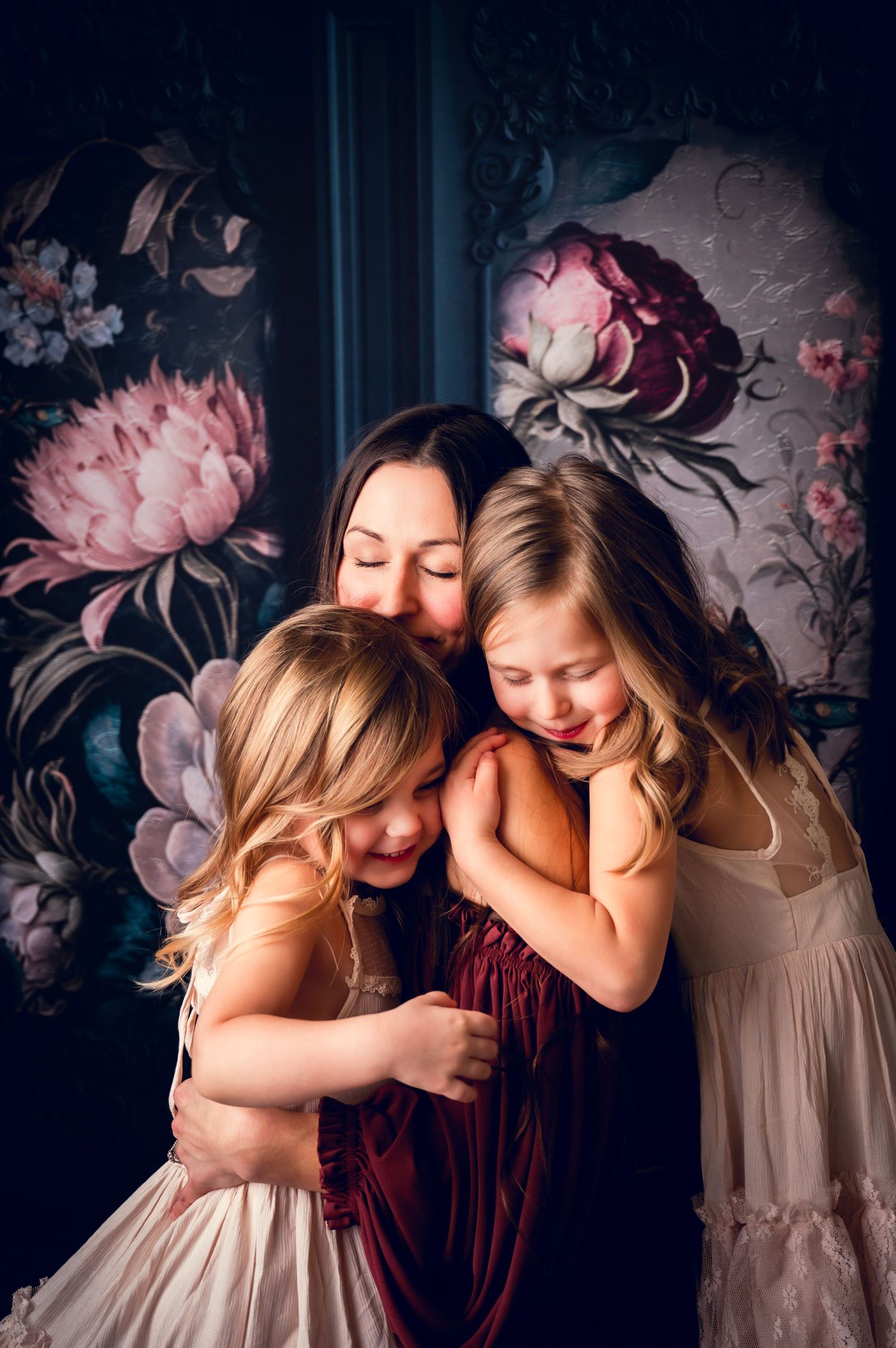 Intimate family portrait with burgundy and cream dresses against dark floral wallpaper.