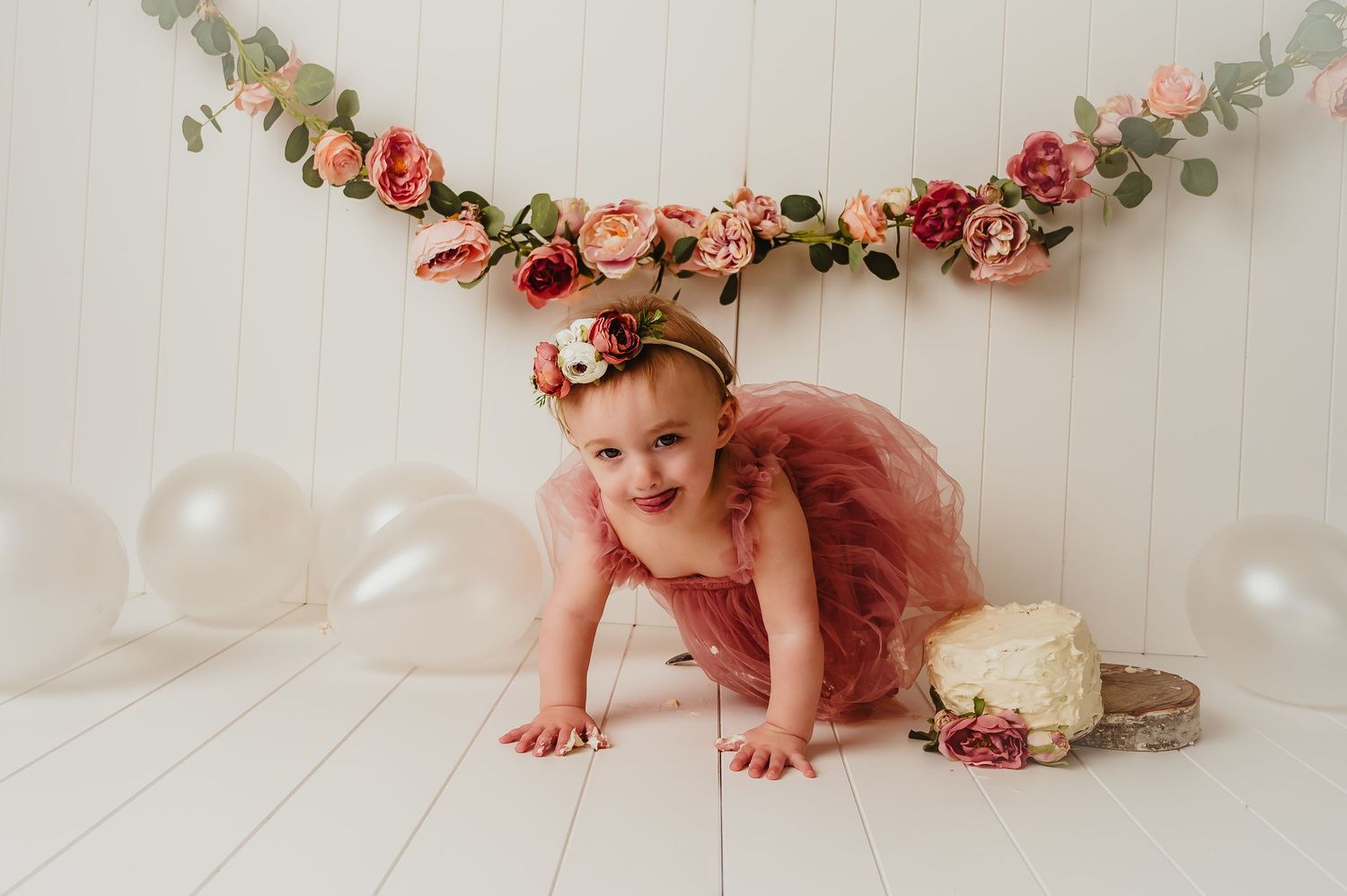 A baby in a pink tulle dress crawls under a floral garland with white balloons in a studio setting.