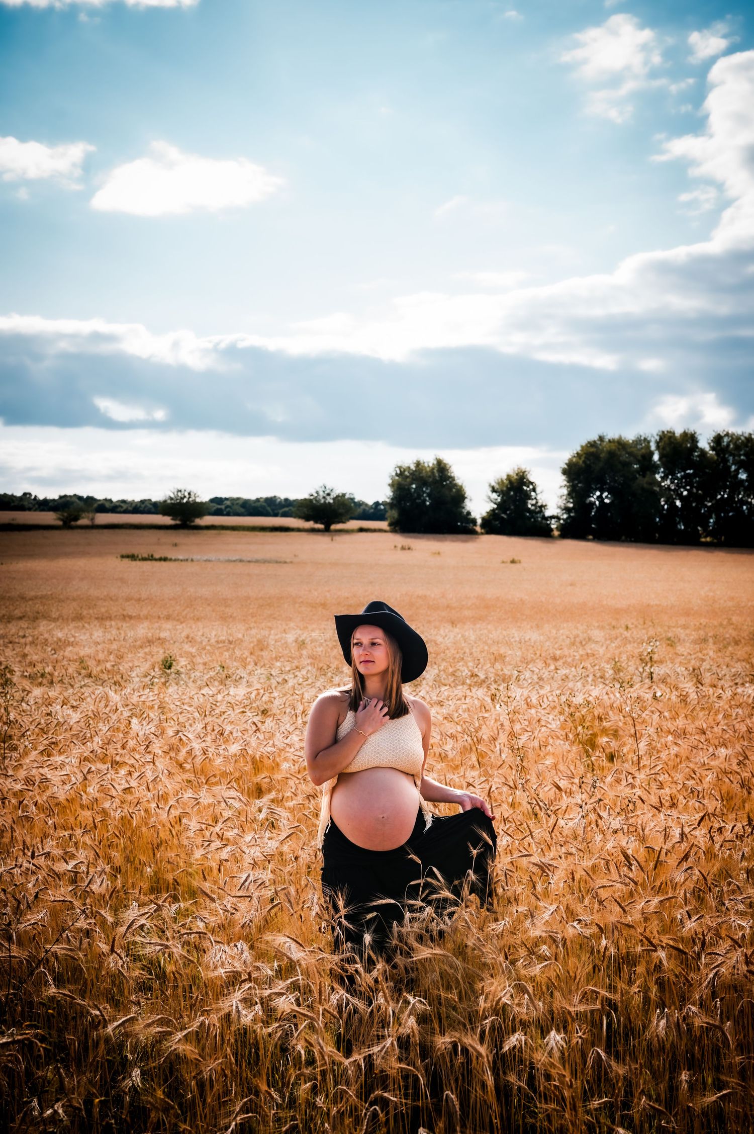 Pregnant woman in black hat cradling her baby bump in a golden wheat field under a partly cloudy sky.