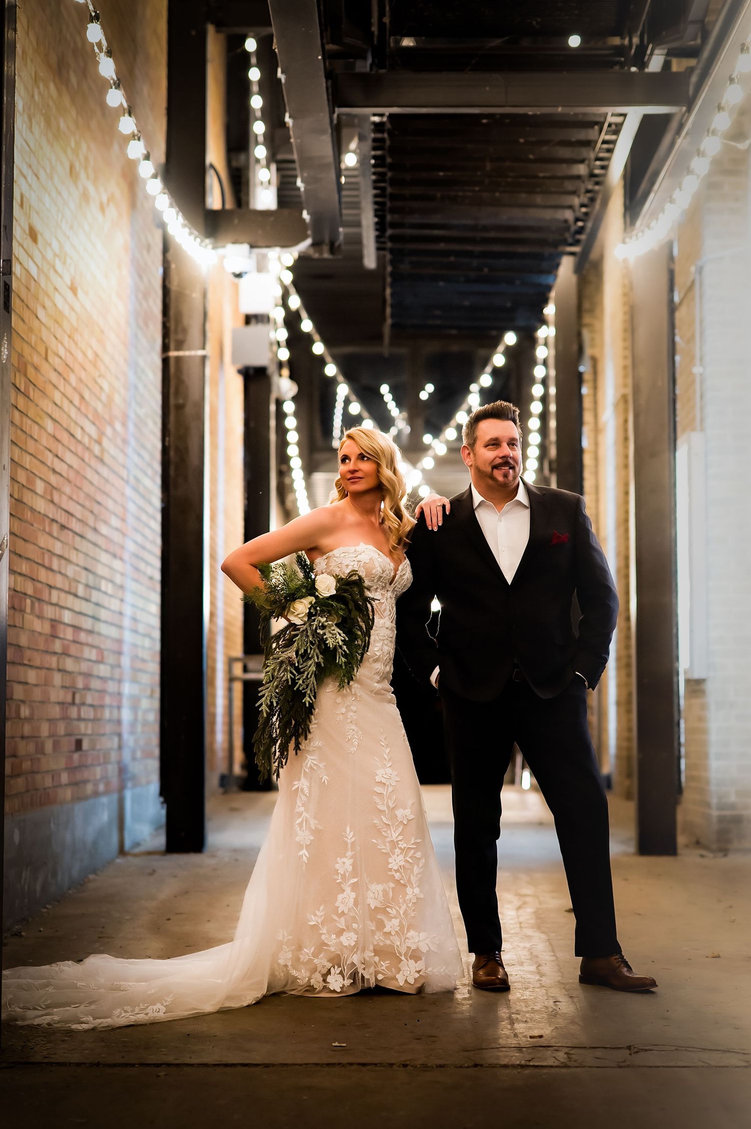 Elegant wedding couple walking through an industrial venue hallway decorated with string lights.